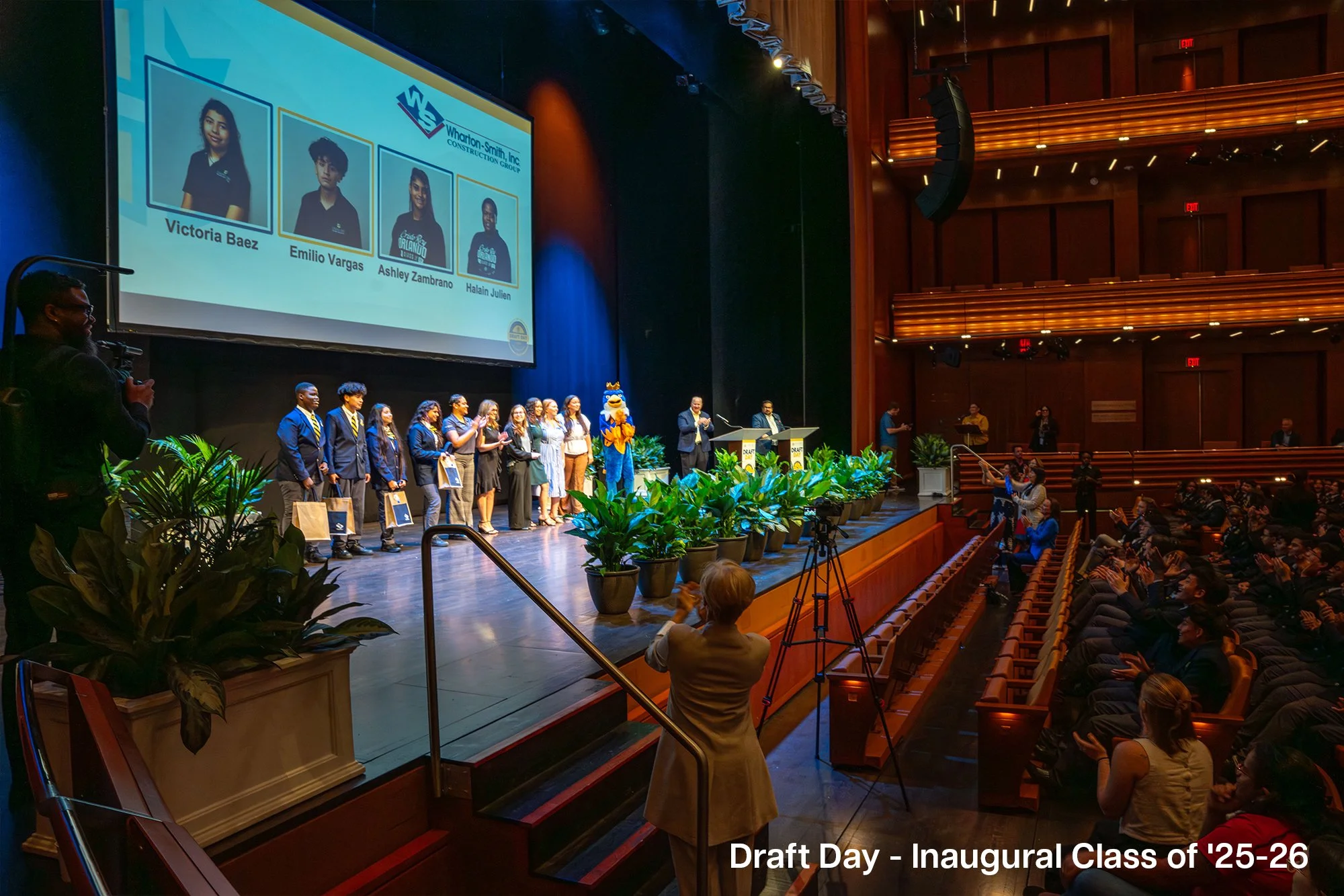 Students on stage receiving awards during the inauguration of the class of 2025-26 on Draft Day, with a large screen behind displaying their names and school logo. Audience applauds, and a woman in the foreground takes a photo.
