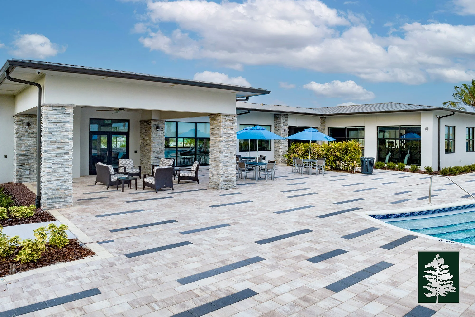 Modern outdoor pool area with patio furniture, umbrellas, and a swimming pool, adjacent to a contemporary building with large windows and stone accents under a partly cloudy sky.