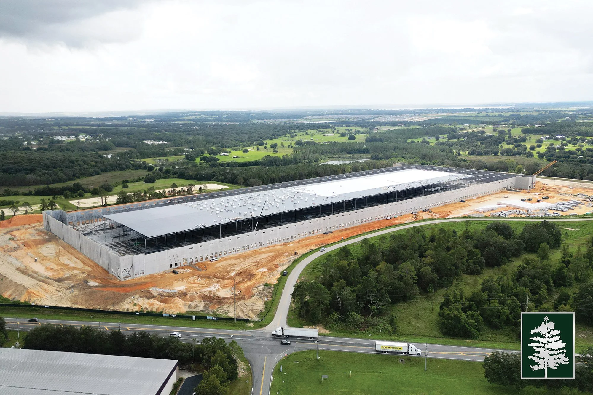 A large building under construction in a semi-rural area with green fields and trees in the background. The building has a metal roof and is surrounded by a dirt site. A road with trucks and cars runs in front of the site. There is a logo of a tree o