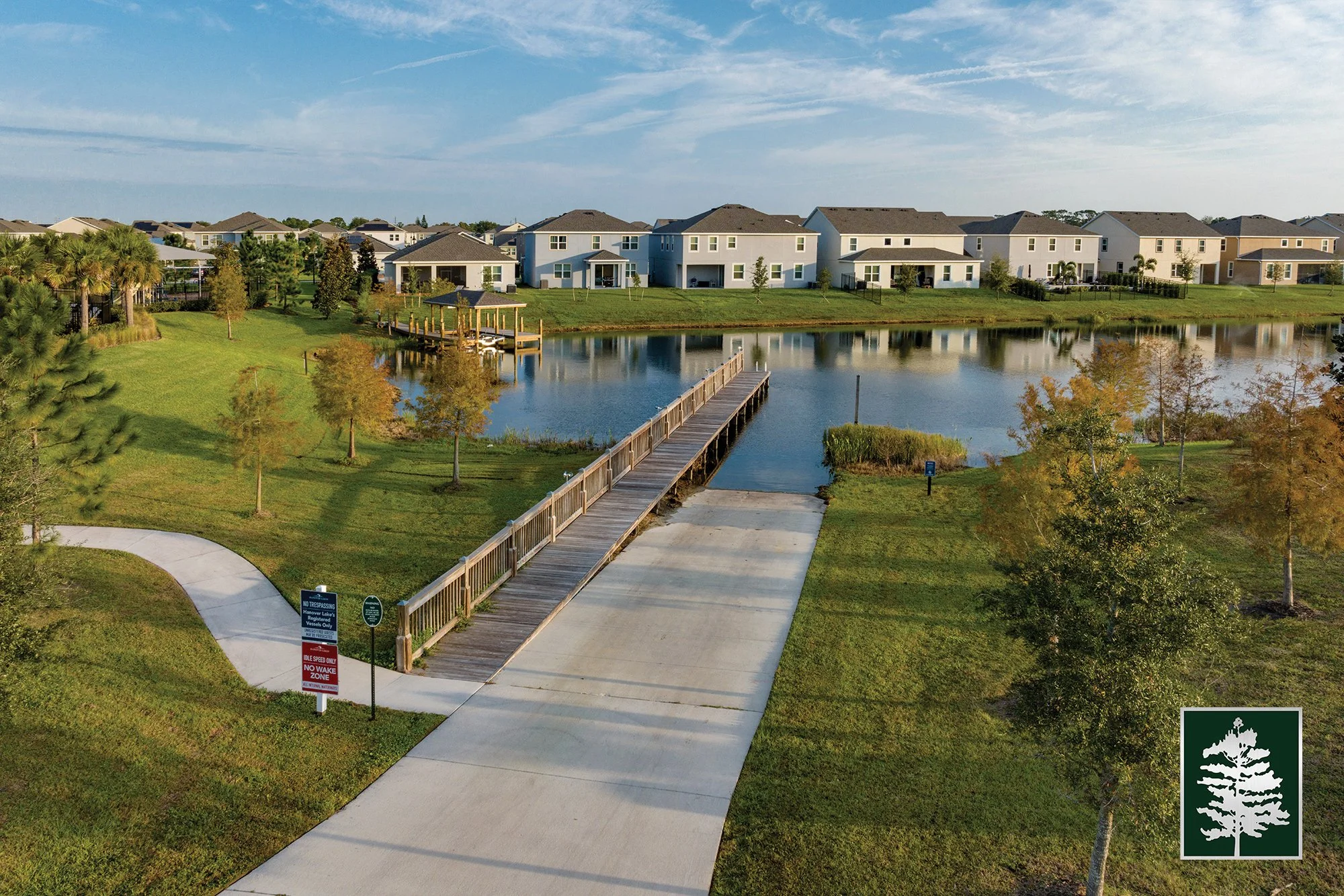 A residential neighborhood with a lake, wooden pier, and modern houses, surrounded by green lawns and trees under a partly cloudy sky.