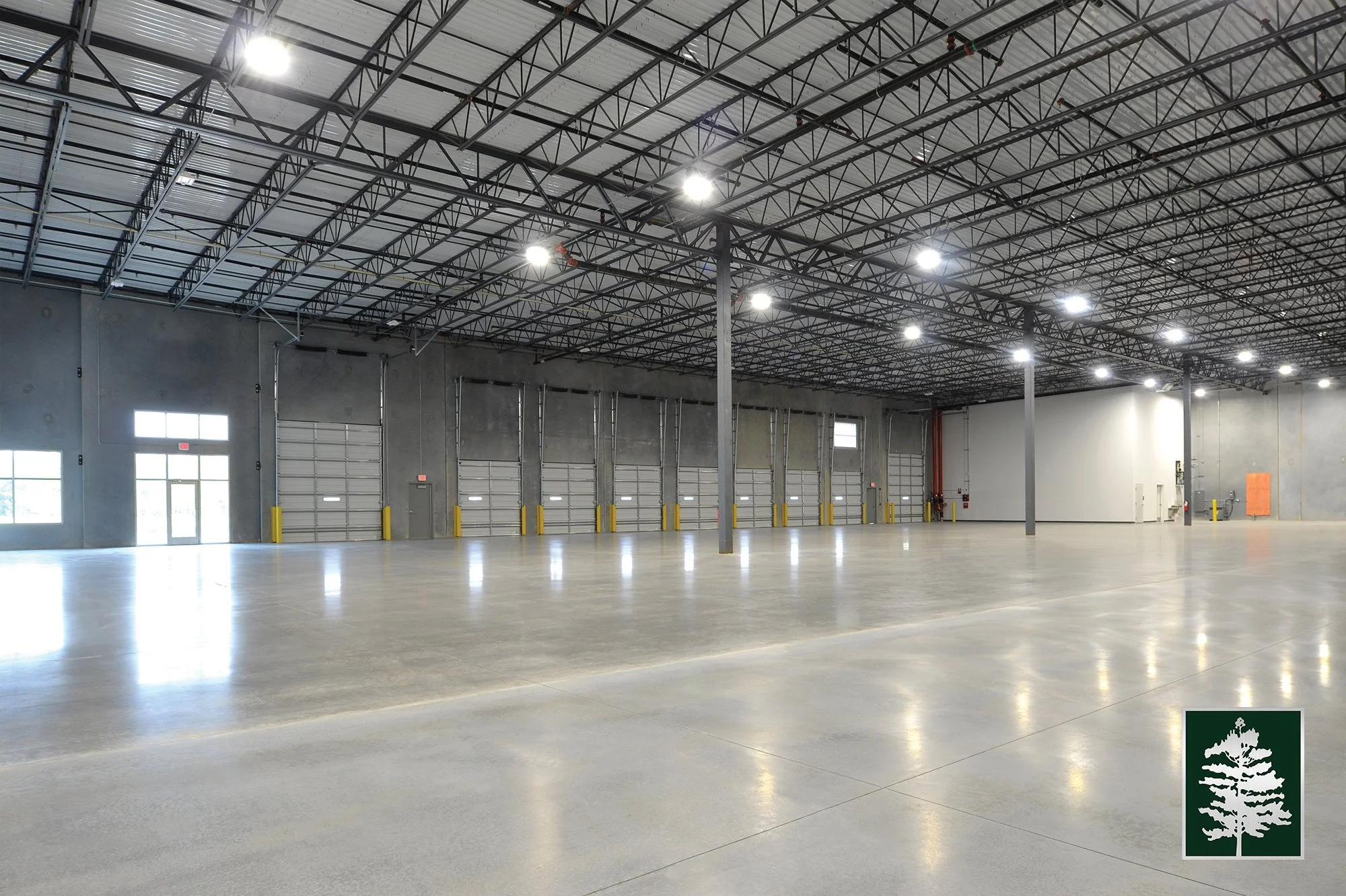 Empty warehouse with high ceiling, concrete floor, multiple large garage doors, and minimal interior features.