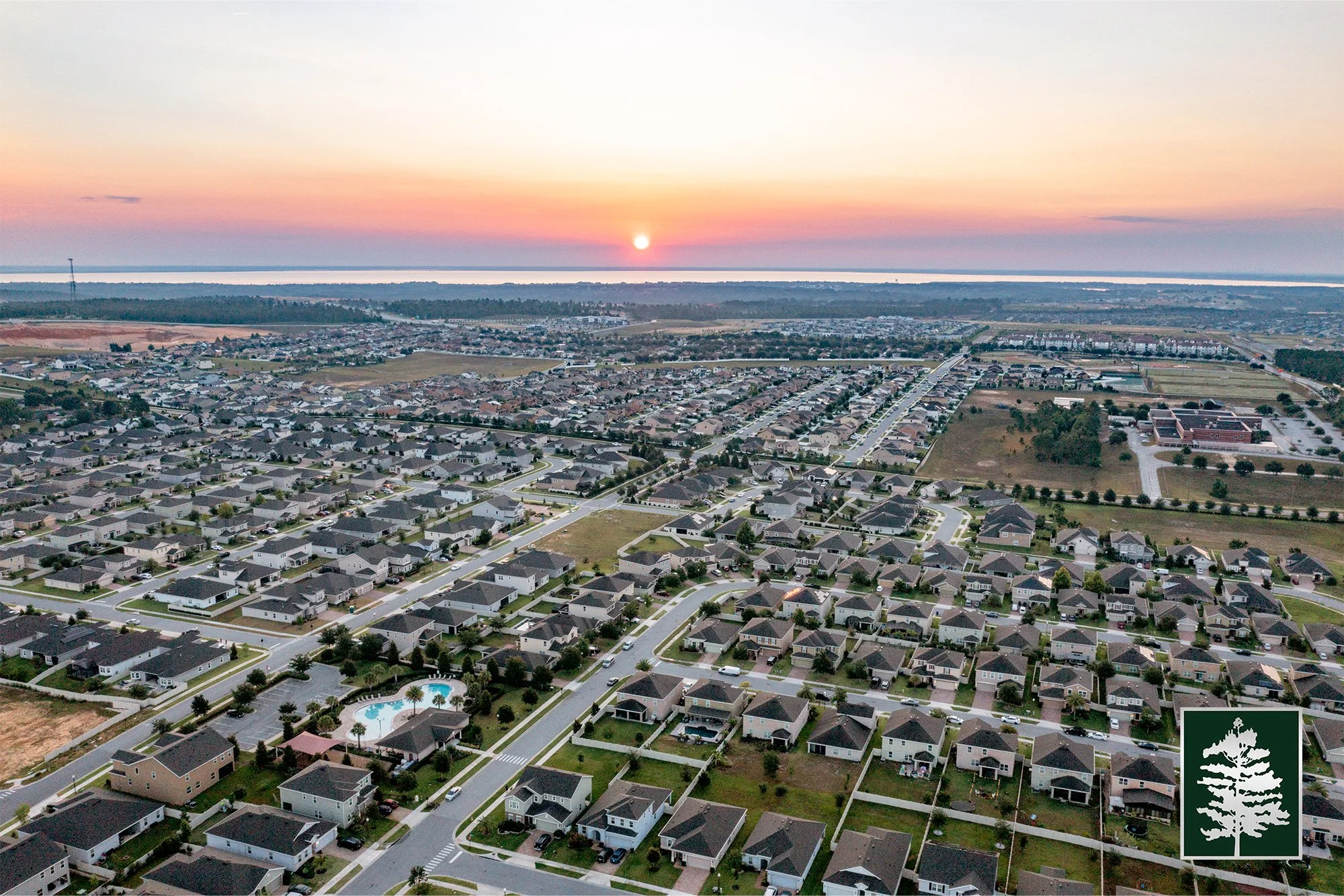 Aerial view of a suburban neighborhood at sunset, showing numerous houses, streets, a swimming pool, and open green spaces.