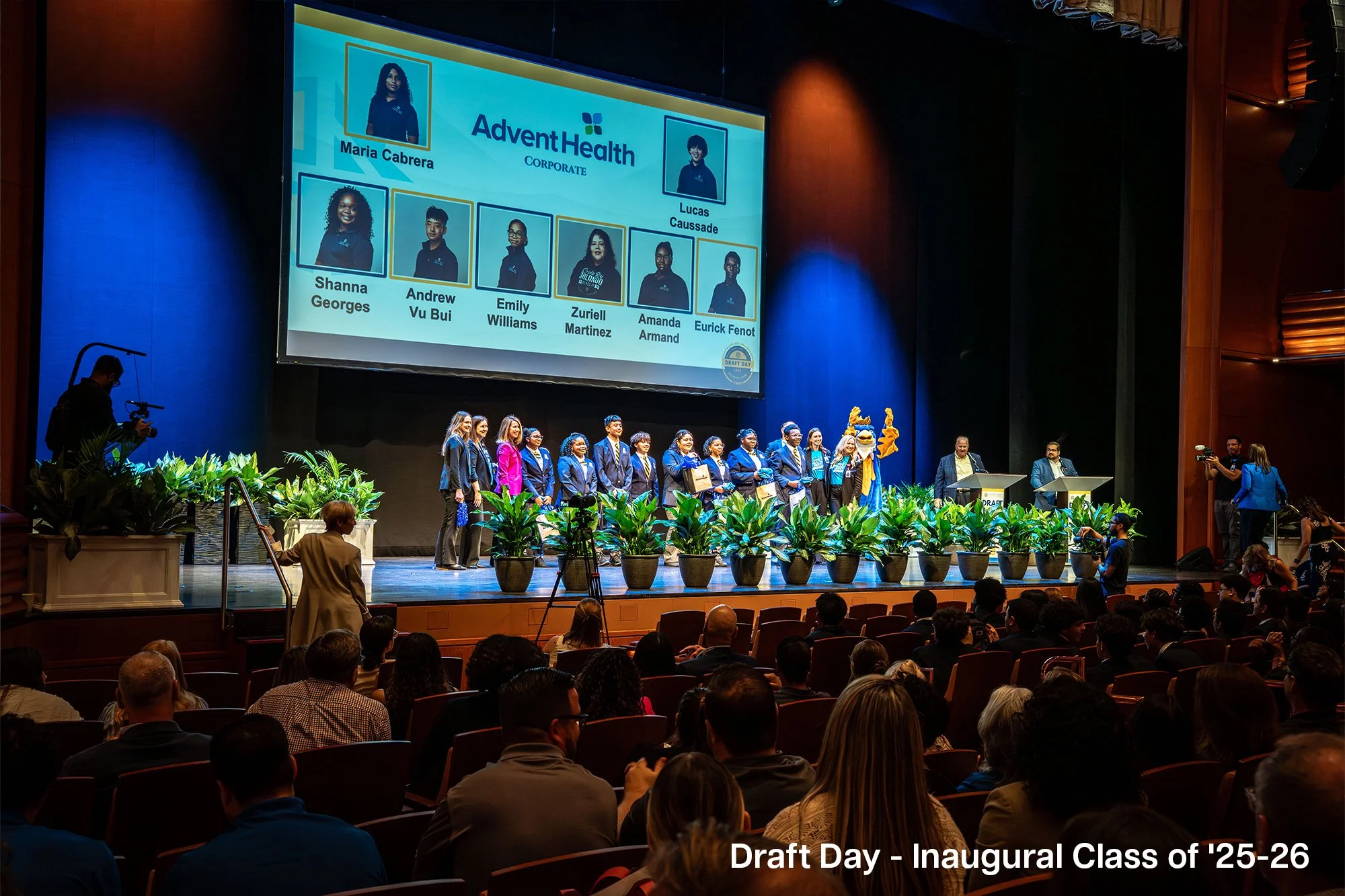 Students on stage accepting awards during a graduation ceremony, with a large screen displaying their names and photos, audience seated in front, and photographers capturing the event, labeled as Draft Day - Inaugural Class of '25-'26.