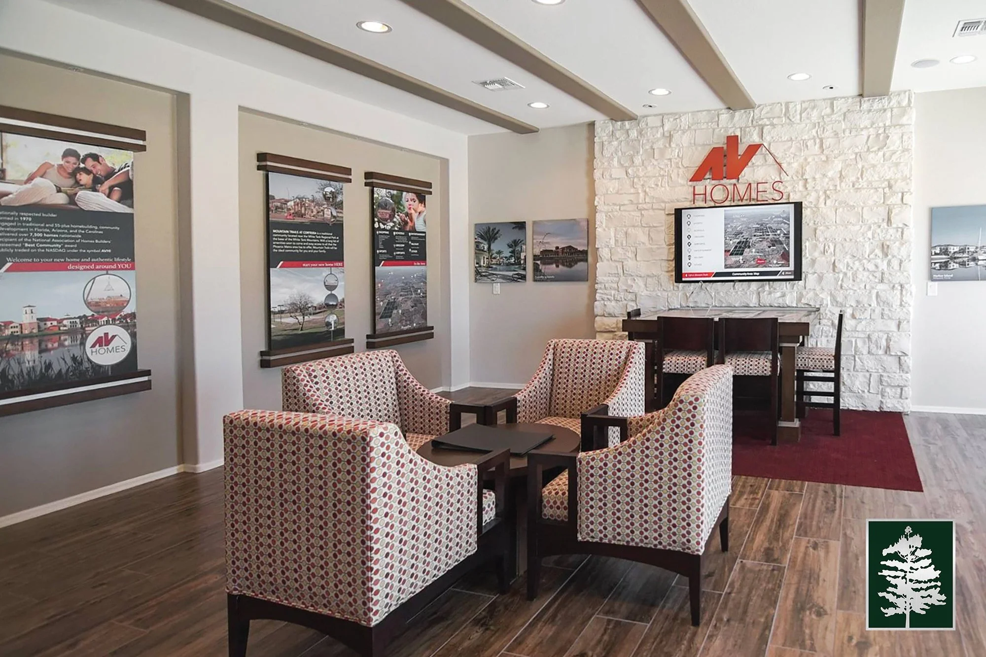 Interior of a real estate office featuring a seating area with four patterned armchairs around a small table, framed promotional posters on the wall, a stone accent wall with a mounted TV displaying real estate listings, and a logo for AV Homes on the wall.