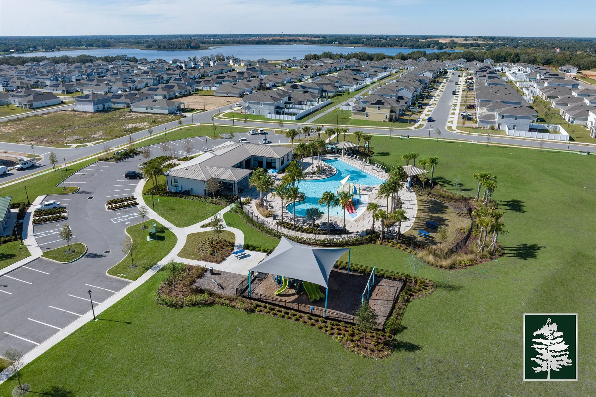 Aerial view of a community pool with palm trees, surrounded by a clubhouse, parking lot, and a neighborhood of houses with paved streets, and a lake in the background.