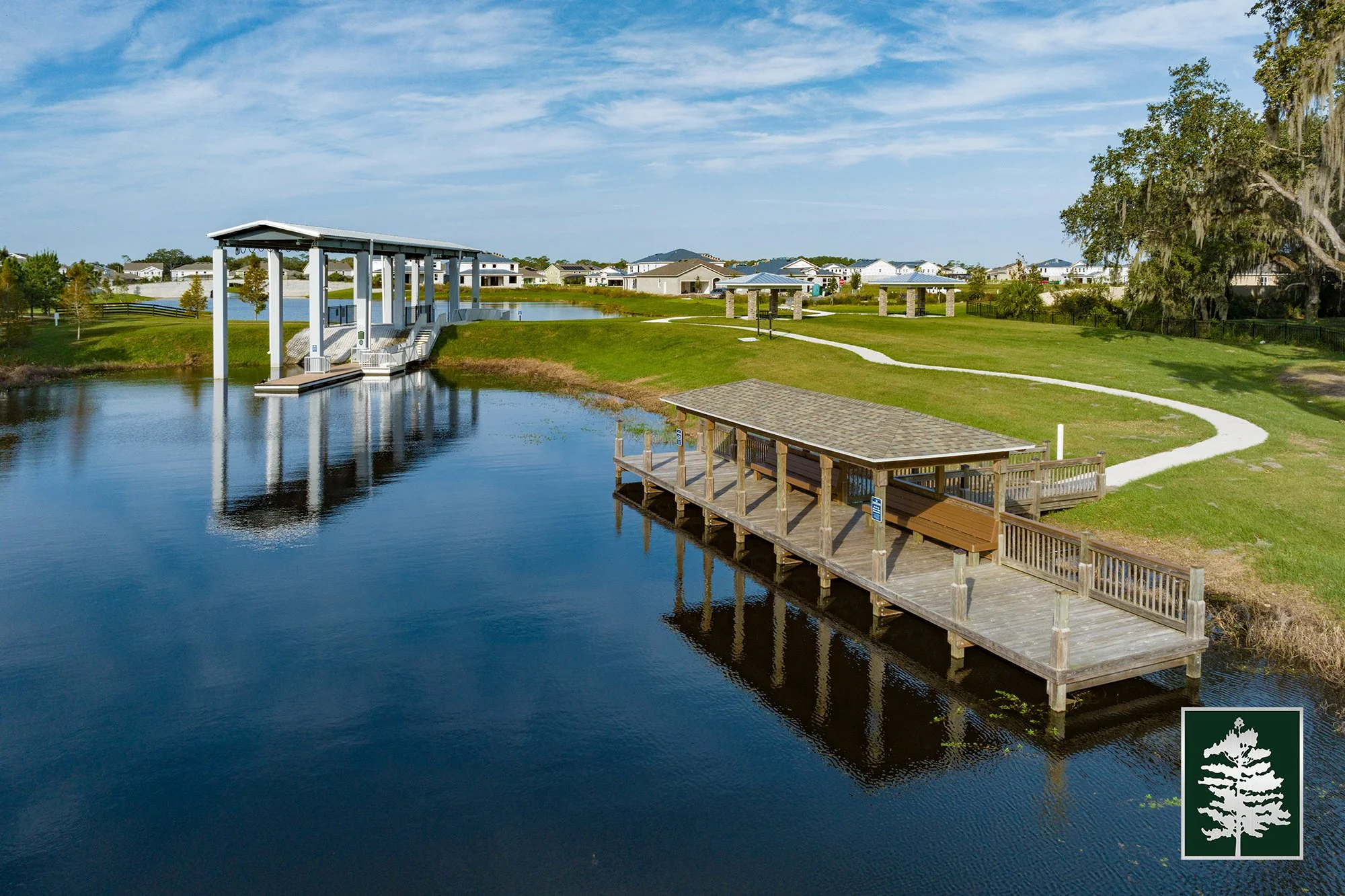 A scenic park with a pond, a covered dock, a walking trail, and residential houses in the background under a partly cloudy sky.