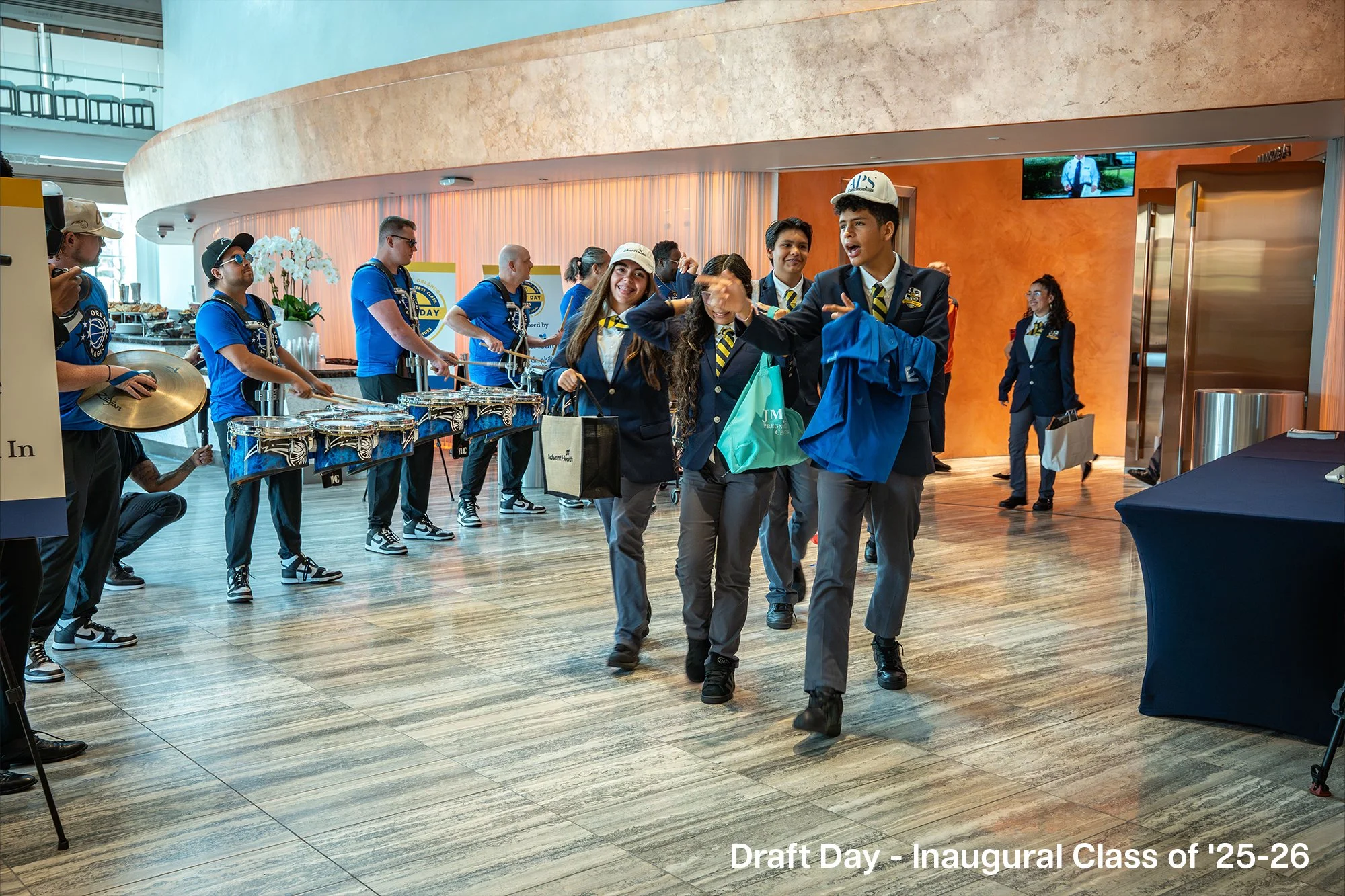 Students in formal attire walk through a lobby, with a band playing percussion instruments in the background, during the Draft Day event for the Inaugural Class of 2025-26.