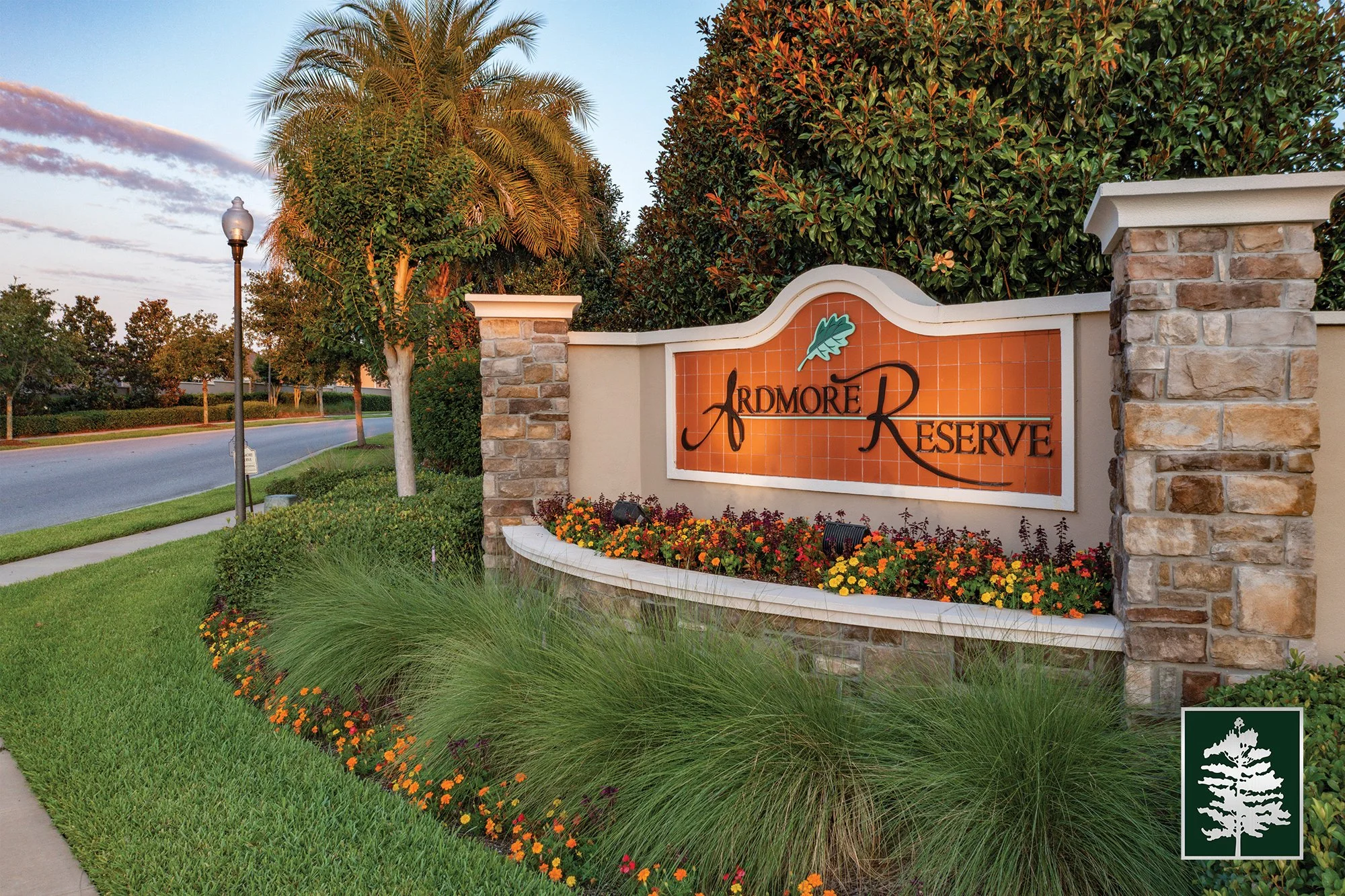 Entrance sign for Ardmore Reserve, with orange tiled background, black text, green leaf decoration, surrounded by flowers and landscaping, with a sidewalk, street, trees, and a lamppost in the background.