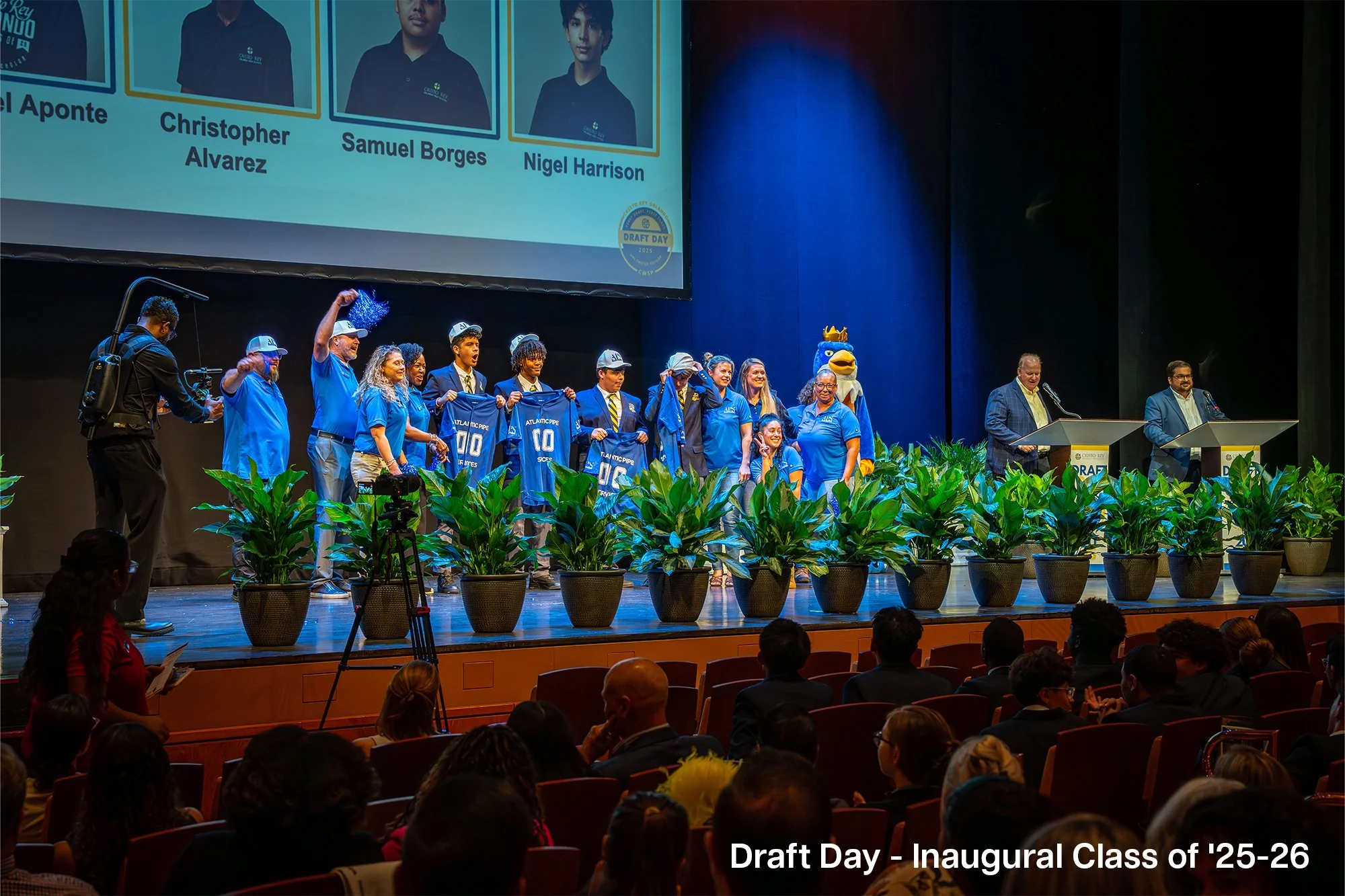 Students on stage during Draft Day event, some holding jerseys, with large screen displaying class photos and names in the background, audience seated in front.