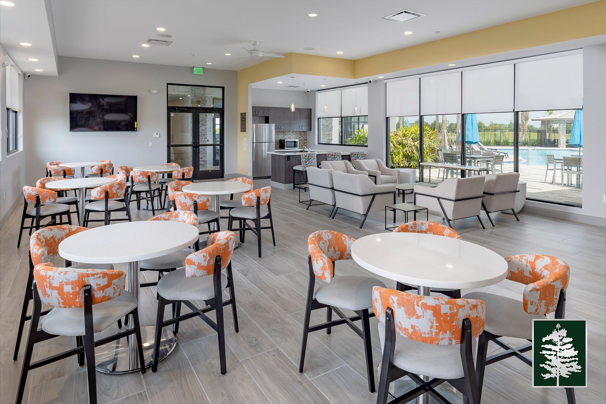 Modern lounge area with round tables and orange patterned chairs, white sofas near large windows overlooking a pool outside, kitchen in the background, and a TV on the gray wall.