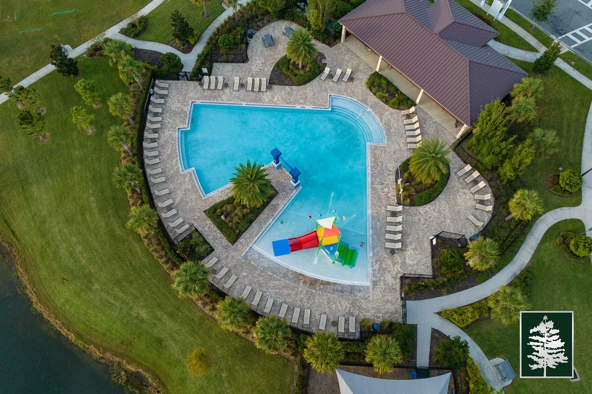 Aerial view of a community swimming pool with surrounding lounge chairs and tropical landscaping, including palm trees, next to a pavilion with a red roof.