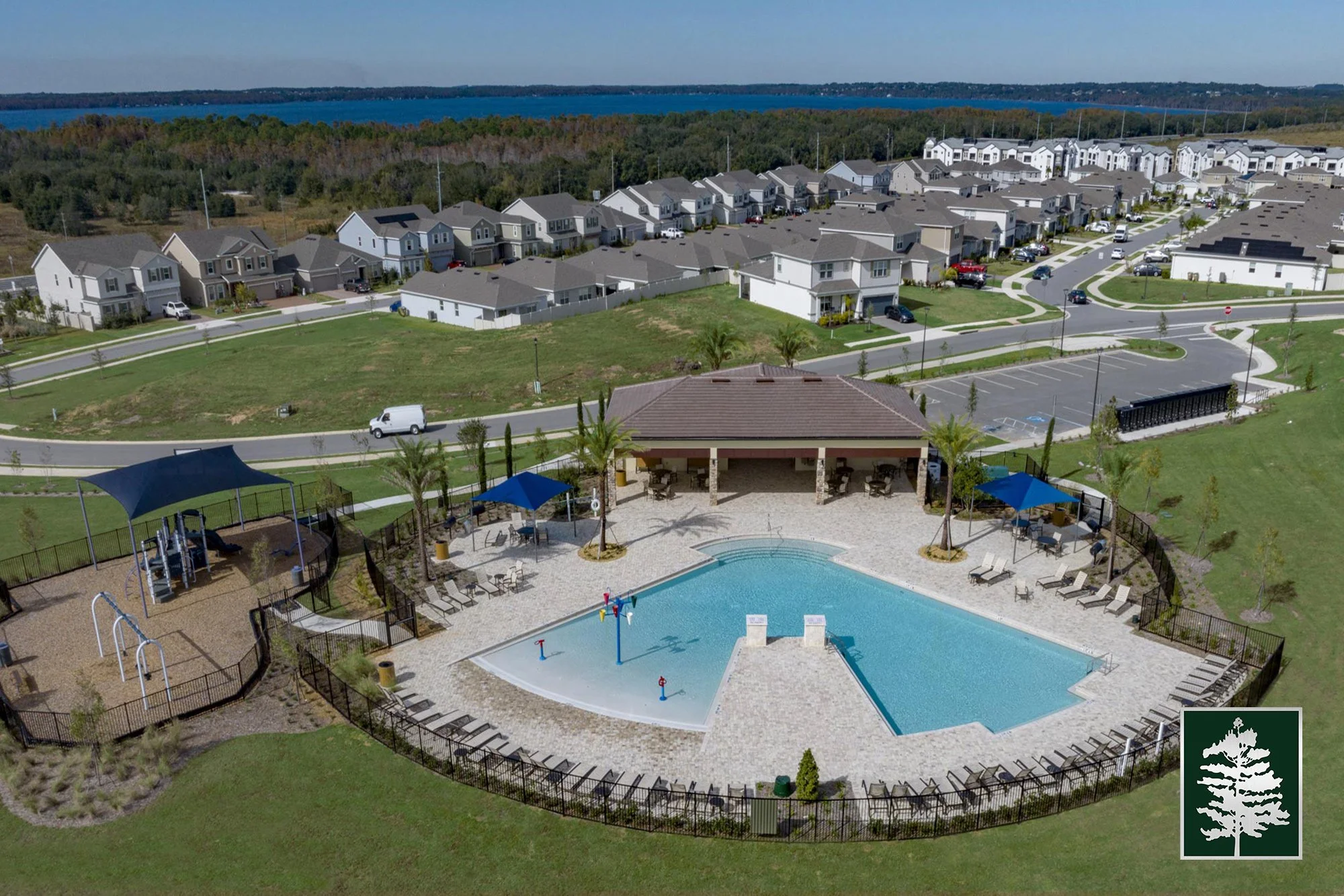 A community swimming pool area with lounge chairs, shade umbrellas, and a water play area, surrounded by a black fence, with a residential neighborhood and a lake in the background.