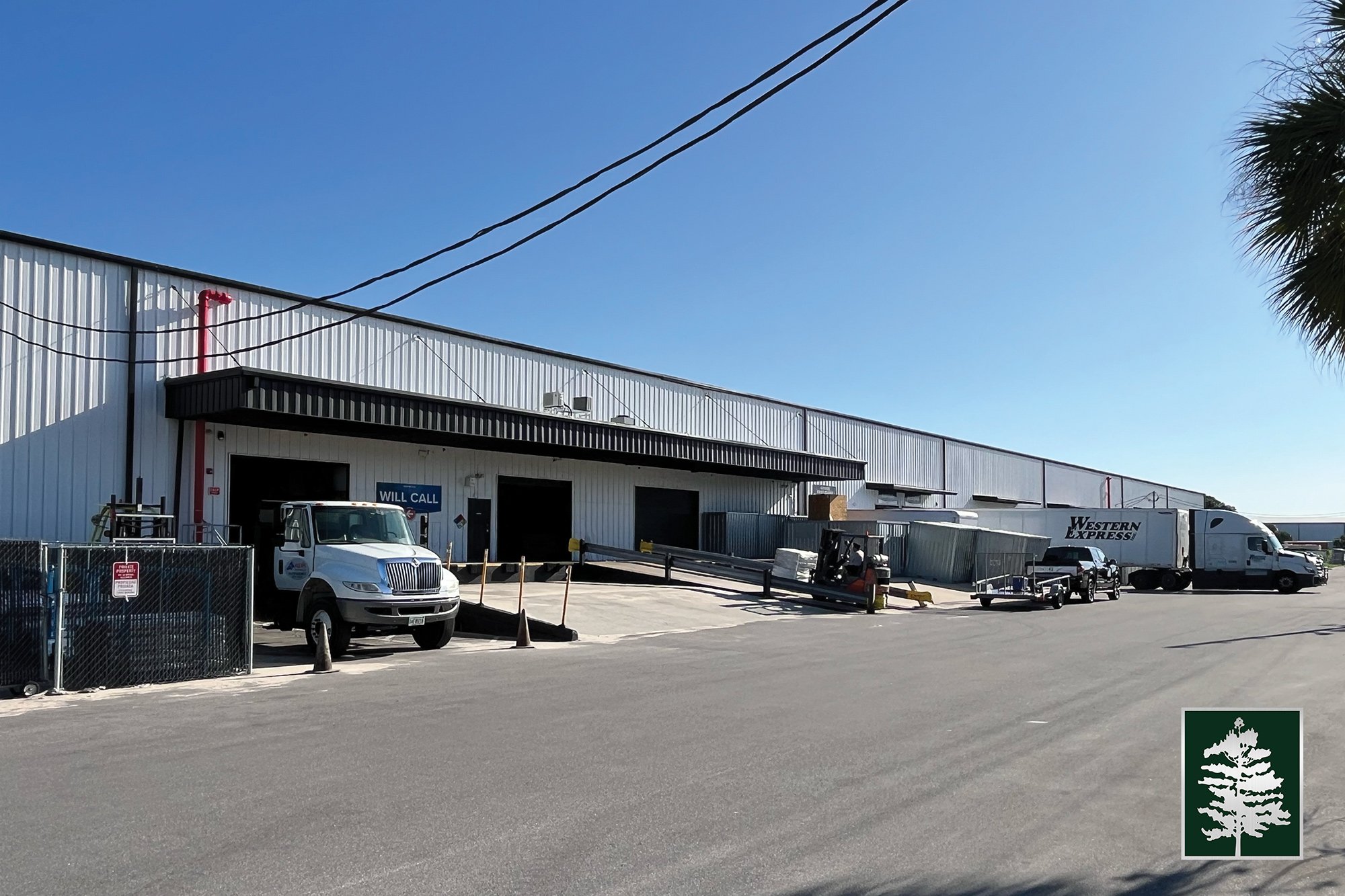Warehouse loading dock with trucks and vehicles, a forklift, and a 'Will Call' sign, under a clear blue sky.