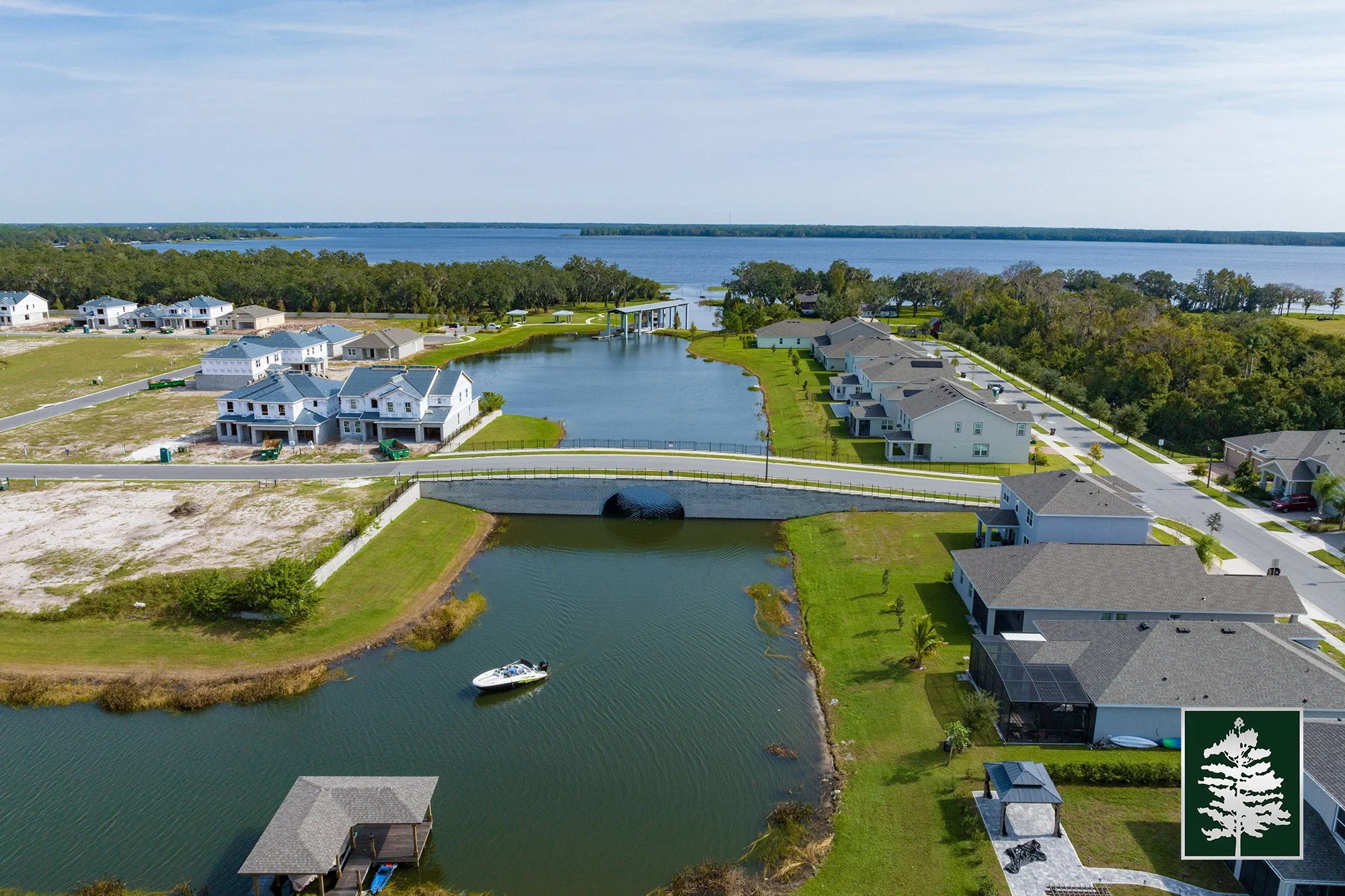 Aerial view of a residential waterfront community with houses lining a canal, a boat in the water, and a bridge crossing over the canal, with a lake in the background.