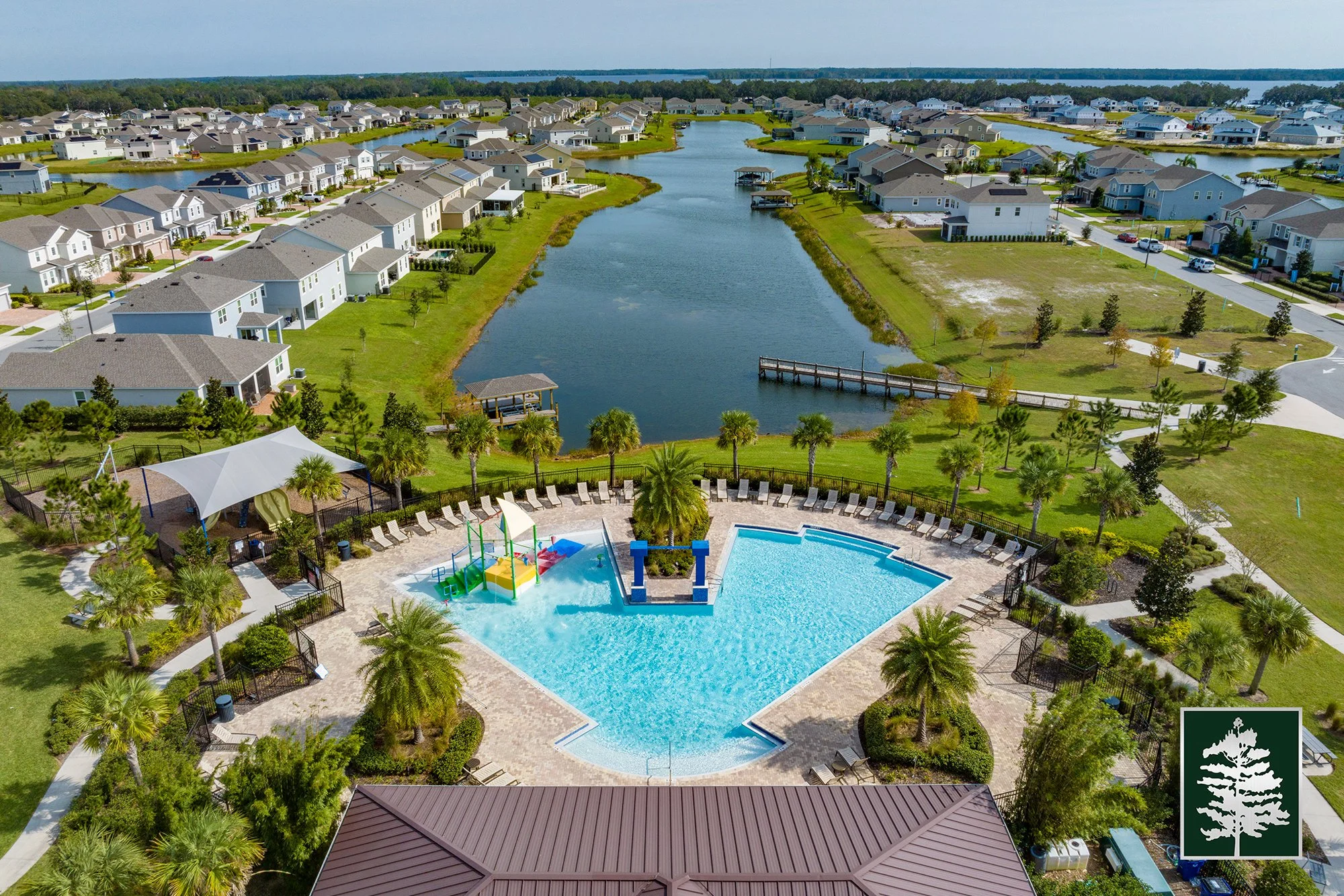 Aerial view of a neighborhood with houses surrounding a waterway, featuring a community pool with a shaded area, palm trees, and a playground with water slides.