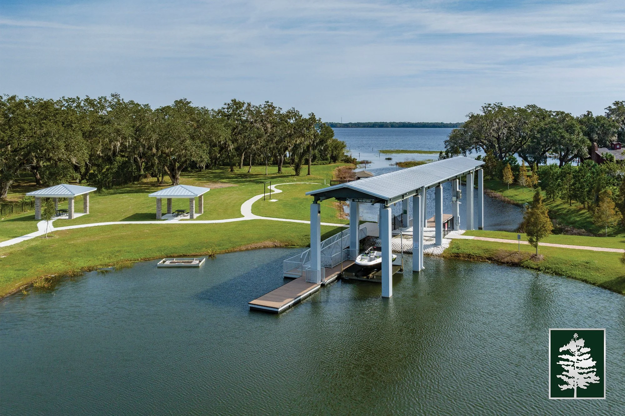 A scenic lakeside park with two pavilions, pathways, and a boat lift with a boat, surrounded by green grass and trees, overlooking the water.