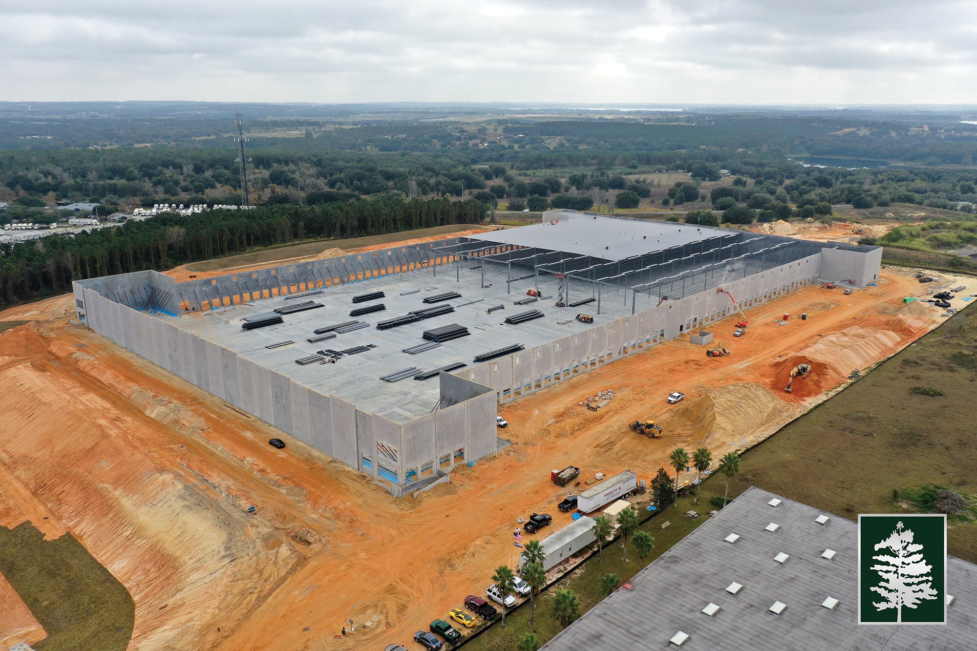 Aerial view of a large building under construction with a concrete foundation and steel framework on a dirt site surrounded by trees and open land.