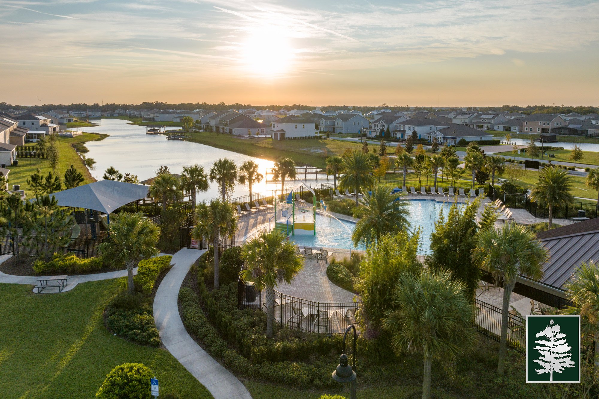 A community swimming pool surrounded by palm trees and lounge chairs, with a playground in the background, overlooking a canal and houses at sunset.
