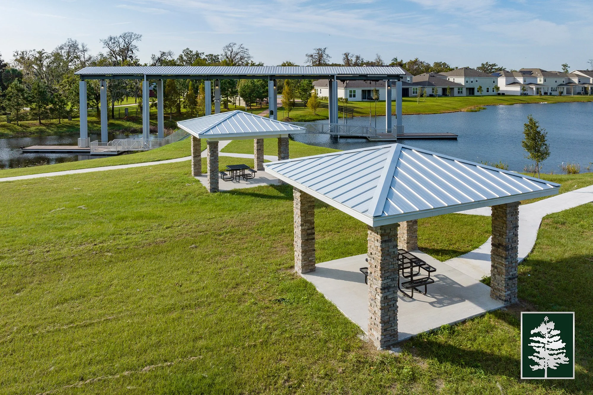 Two covered picnic shelters with brick pillars and metal roofs in a park near a body of water. The park has green grass, walking paths, and trees. In the background, there is a lake with a walking bridge and residential houses.
