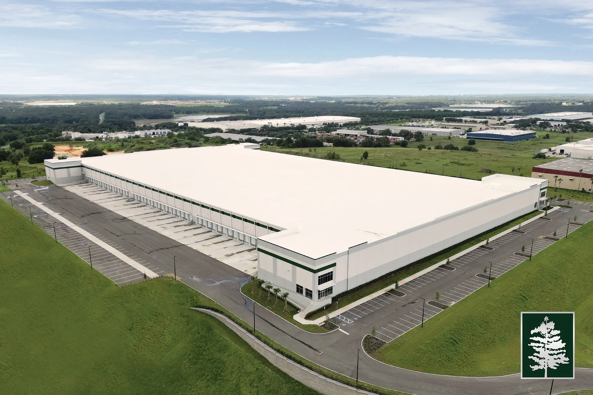 Large white industrial warehouse building with adjacent parking lot surrounded by green grass and a scenic landscape with additional industrial buildings in the background.