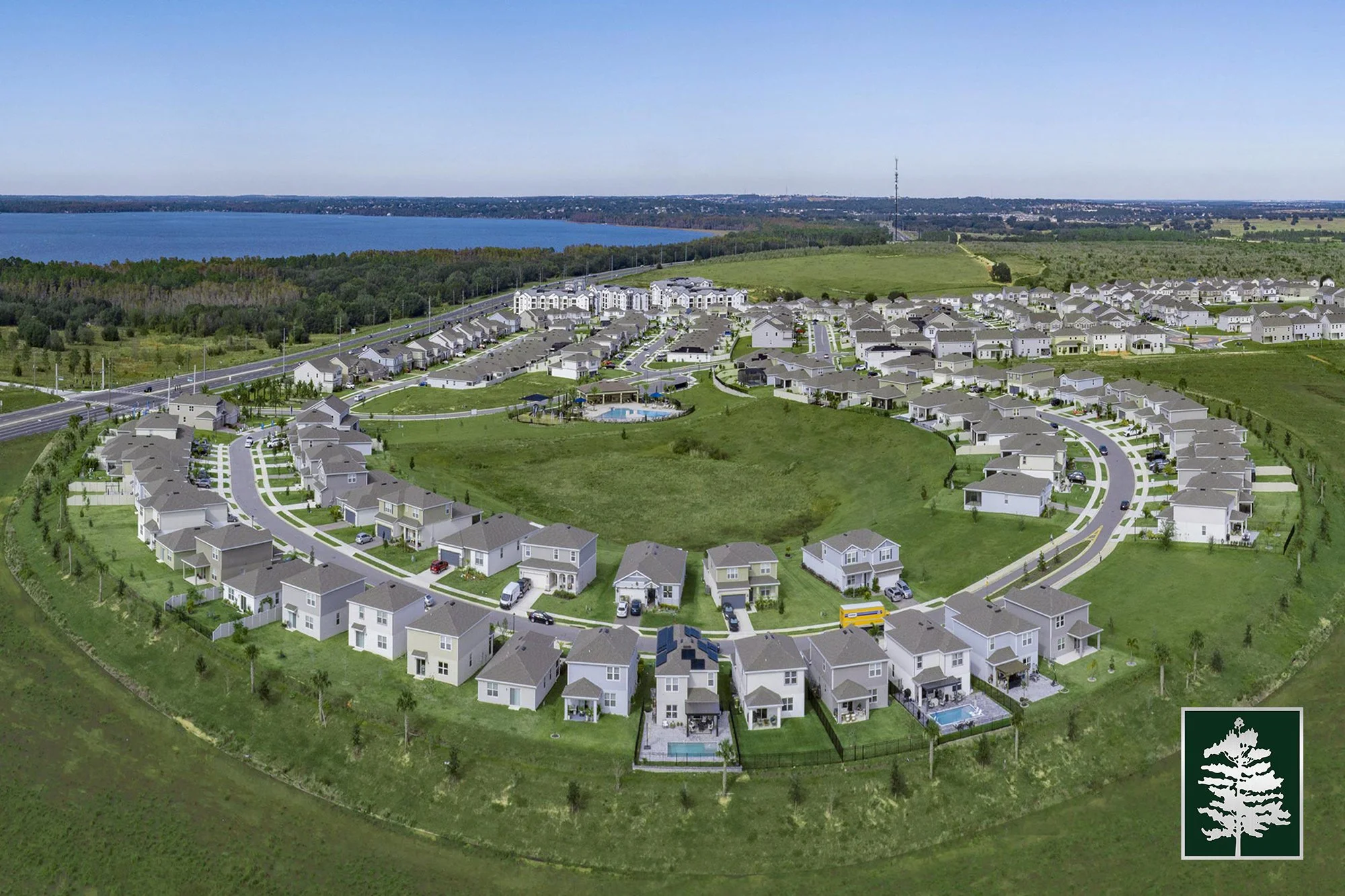 Aerial view of a residential community with houses arranged in concentric circles around a central green space with a pool. Houses have front lawns and driveways. In the background, there is a large body of water, forests, and open fields.