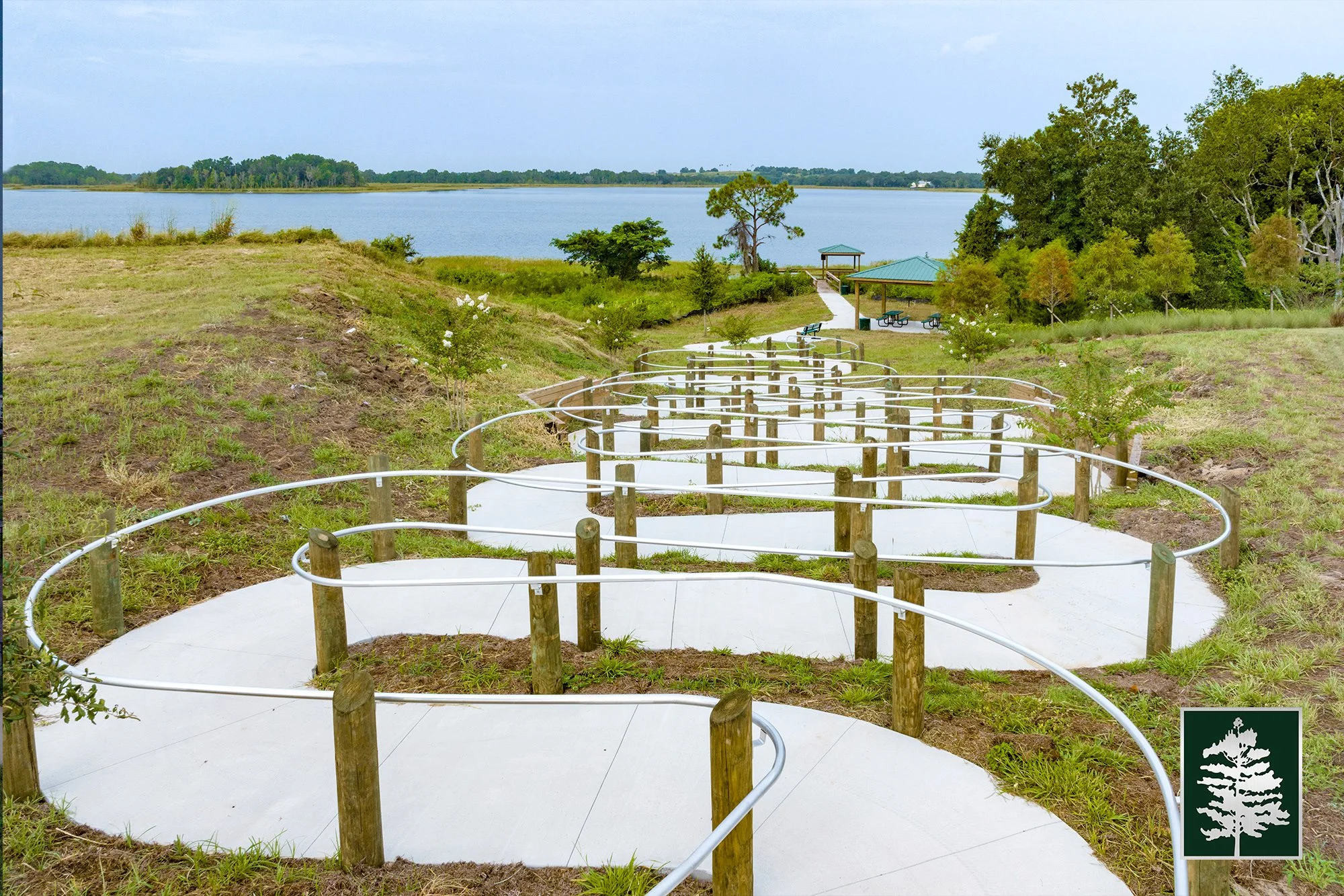 A winding concrete pathway with metal handrails in a park near a body of water, with trees and grass along the sides and a pavilion in the background.