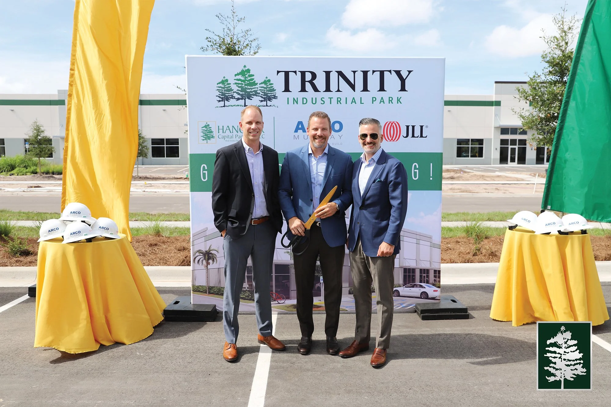Three men in suits standing together for a photo at a groundbreaking ceremony for Trinity Industrial Park, with a backdrop featuring the park's logo and a building. Two tables with yellow tablecloths holding white hard hats with the Arco logo are on 