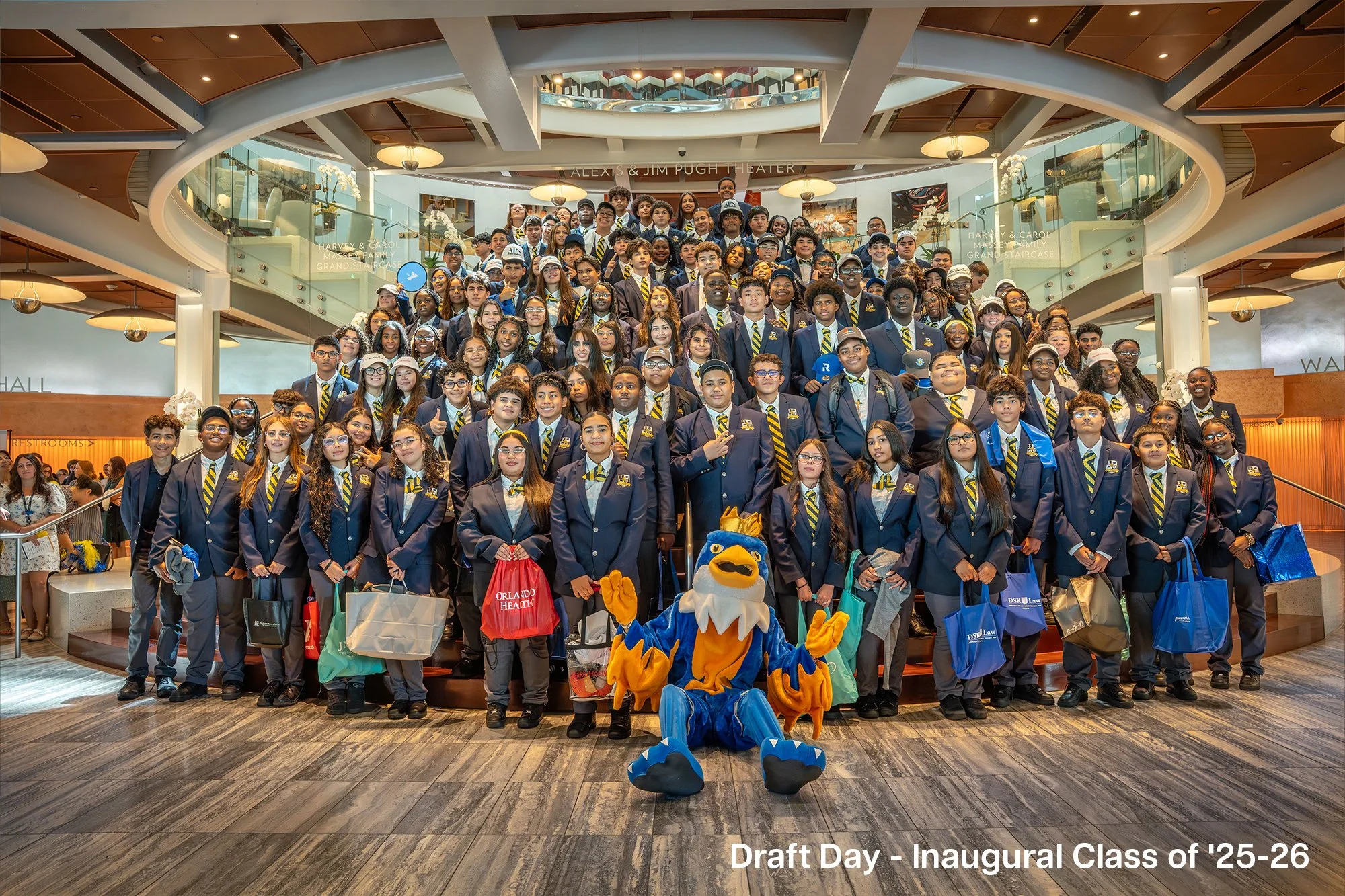 Group of students in blue uniforms with yellow and blue striped ties, posing for a group photo on a staircase inside a building. There is a mascot sitting in front of them. The caption reads 'Draft Day - Inaugural Class of '25-26'.