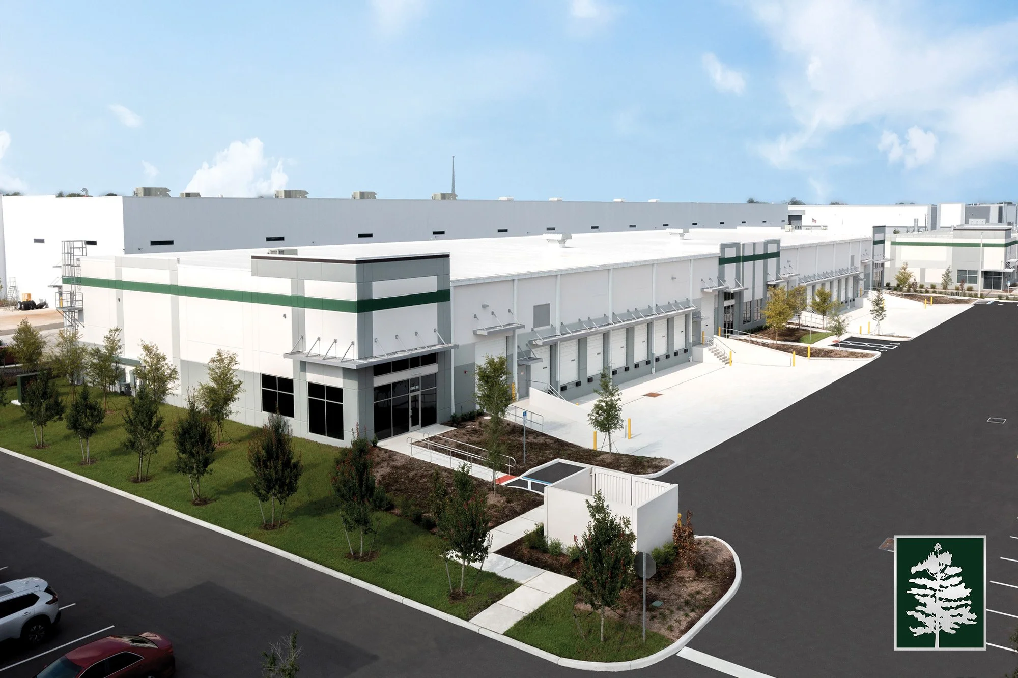 Large modern white warehouse with loading docks, surrounded by sidewalks, green lawns, trees, parking lot, and a clear blue sky.