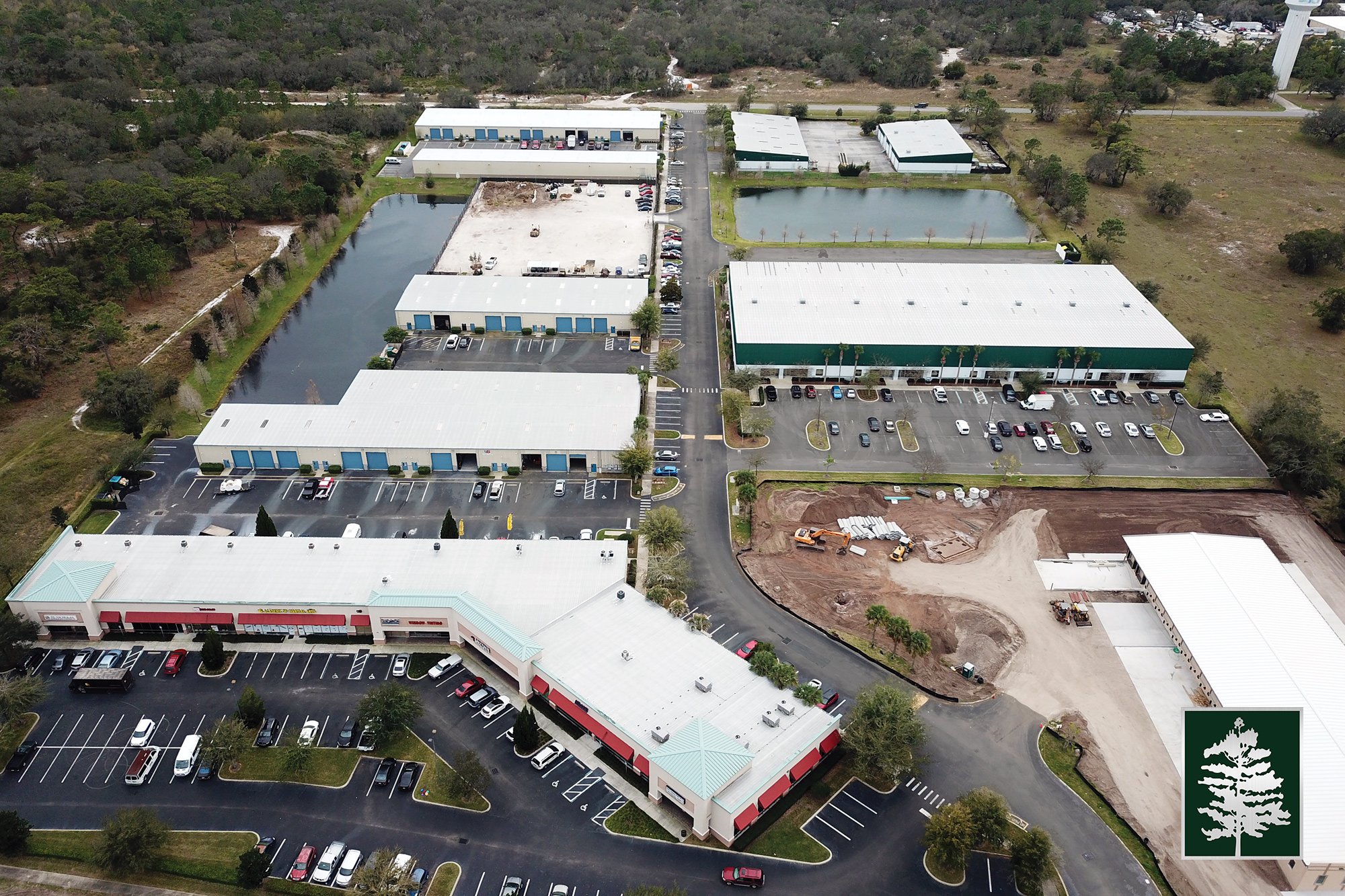 Aerial view of a commercial complex with multiple buildings, parking lots, and construction areas amid trees and greenery.