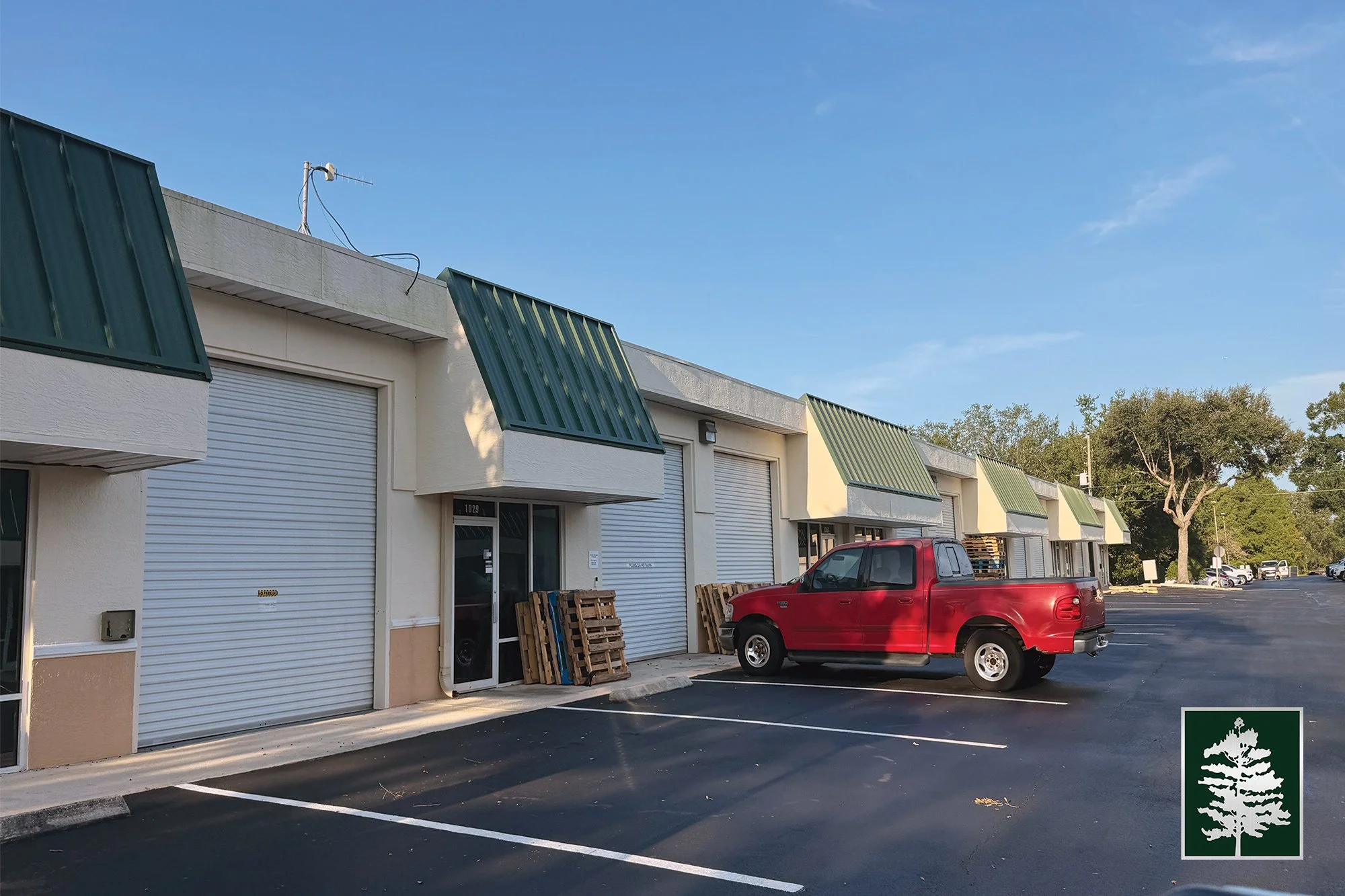 A row of commercial buildings with green metal awnings, white walls, and roll-up garage doors, one with a red pickup truck parked outside, parking lot in front, with trees in the background and a clear blue sky.