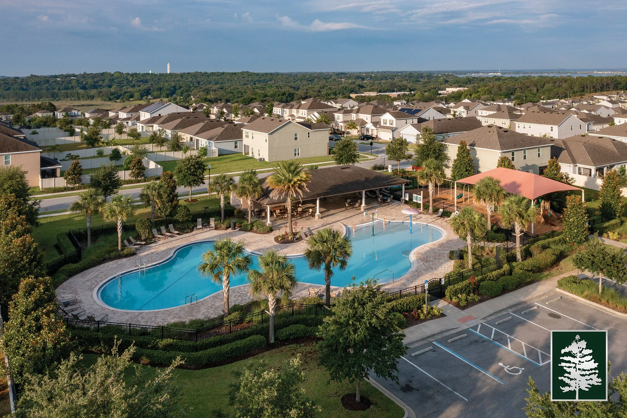 Aerial view of a community swimming pool area with palm trees, lounge chairs, and shaded seating, surrounded by a parking lot and residential houses in the background.