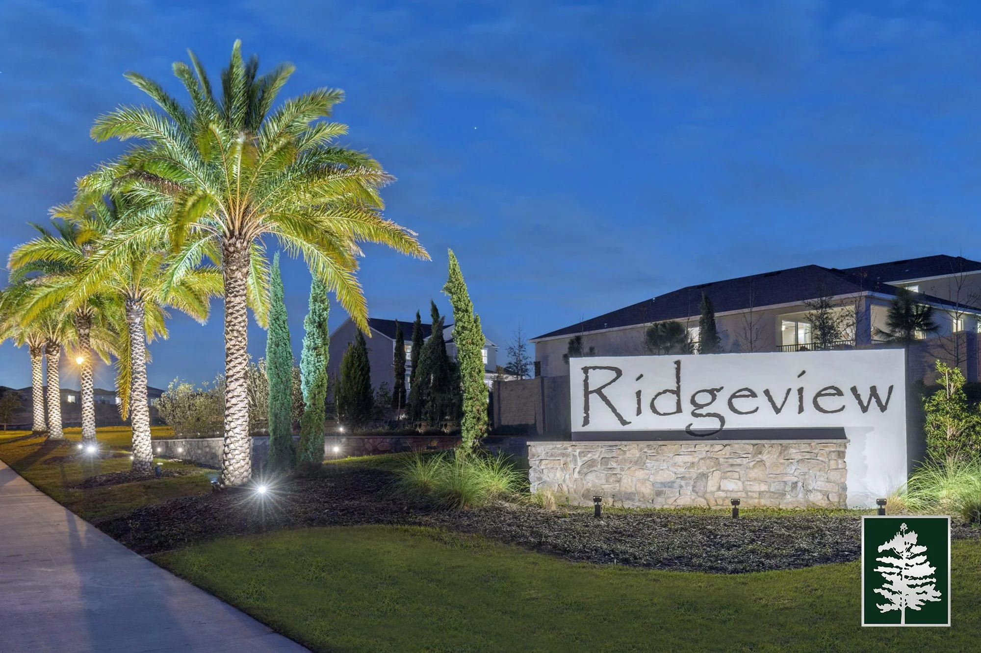 Night view of a neighborhood entrance with a large white sign reading 'Ridgeview' in front of a stone base, surrounded by palm trees and other greenery, with houses in the background and illuminated by landscape lighting.