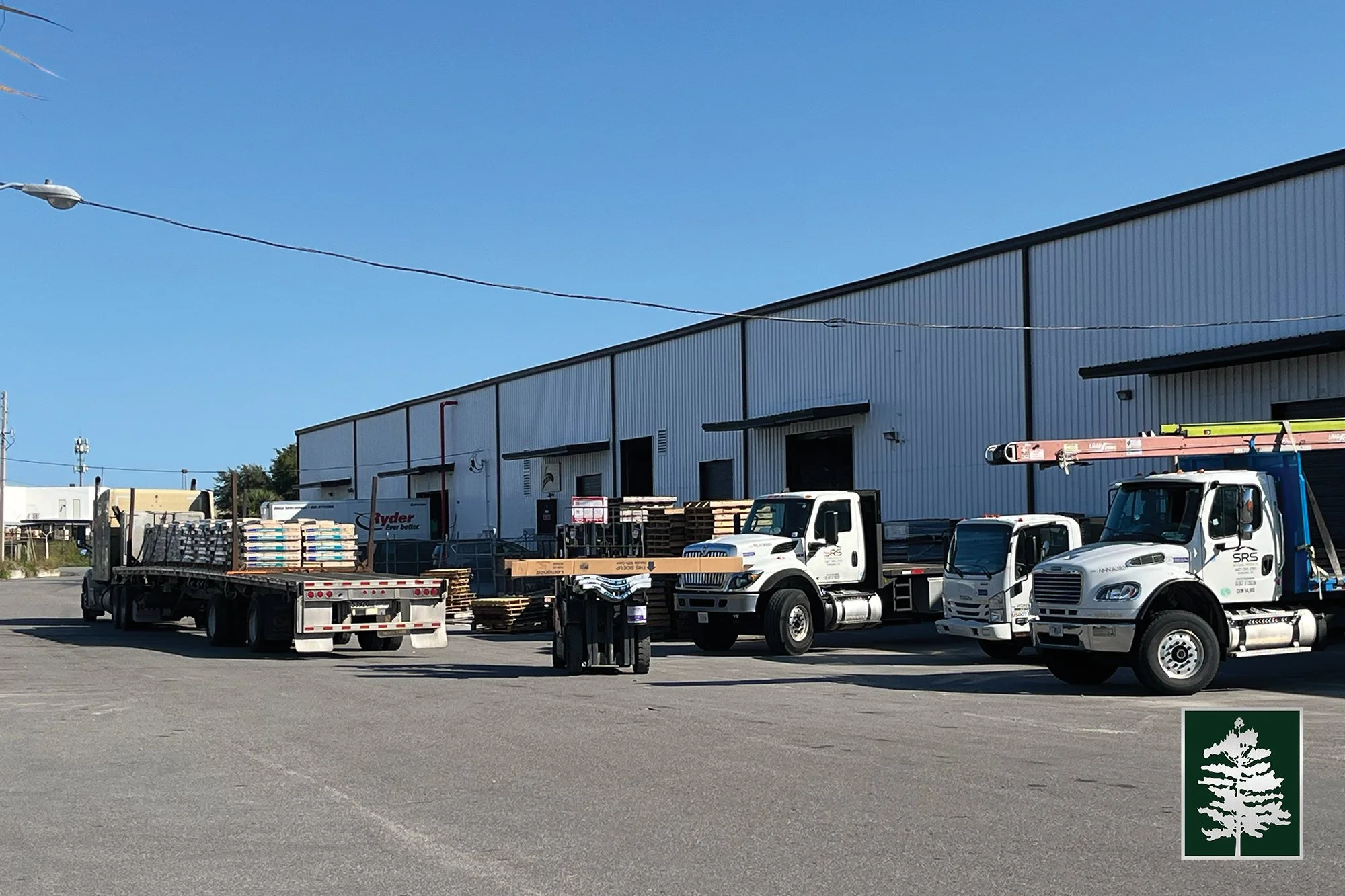 Loading dock area with trucks parked outside a large industrial warehouse, with pallets and bags of materials ready for transportation, under a clear blue sky.