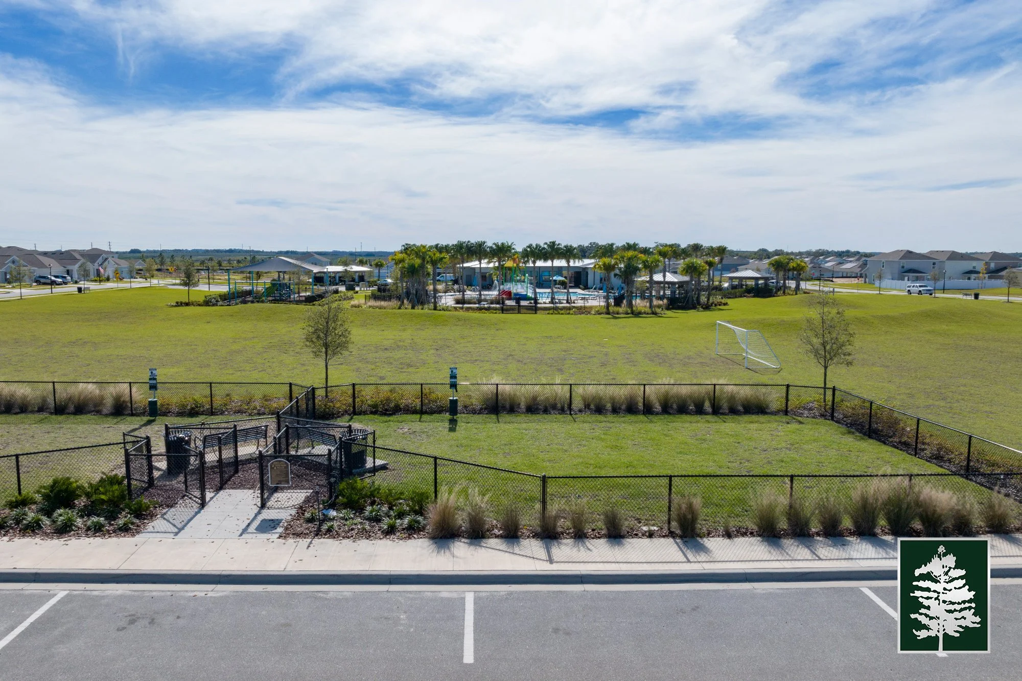 A playground, soccer goal, and picnic area in a community park with a parking lot and residential houses in the background under a partly cloudy sky.