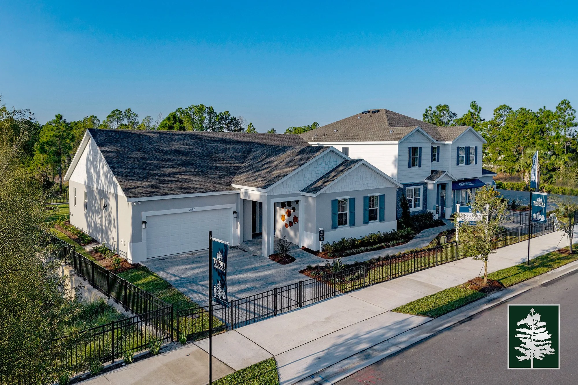 A modern white house with a two-story section and attached garage, surrounded by a black metal fence and landscaped yard with small trees and shrubs, located on a suburban street under a clear blue sky.