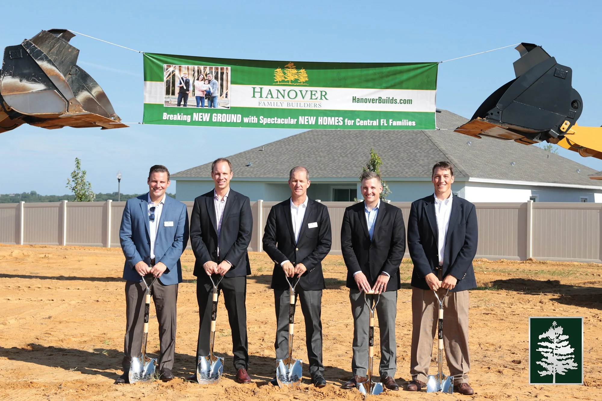 Five men in suits participating in a ground-breaking ceremony with shovels. They are standing on dirt in front of a house with a fenced yard. There is a green banner overhead promoting new homes for Central Florida families. Construction equipment is visible above, and a small logo with a tree symbol is in the lower right corner.