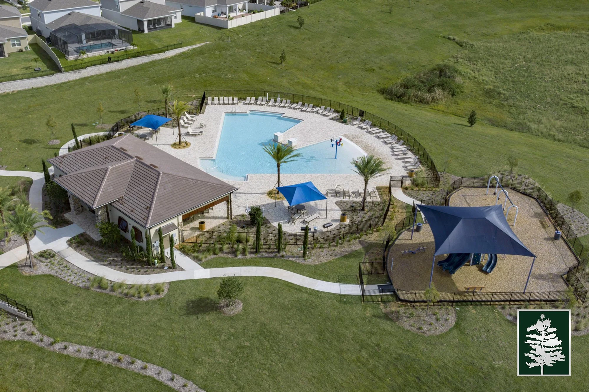 Aerial view of a community pool area with lounge chairs, umbrellas, palm trees, a small building, and a playground, surrounded by a grassy landscape and neighboring houses.
