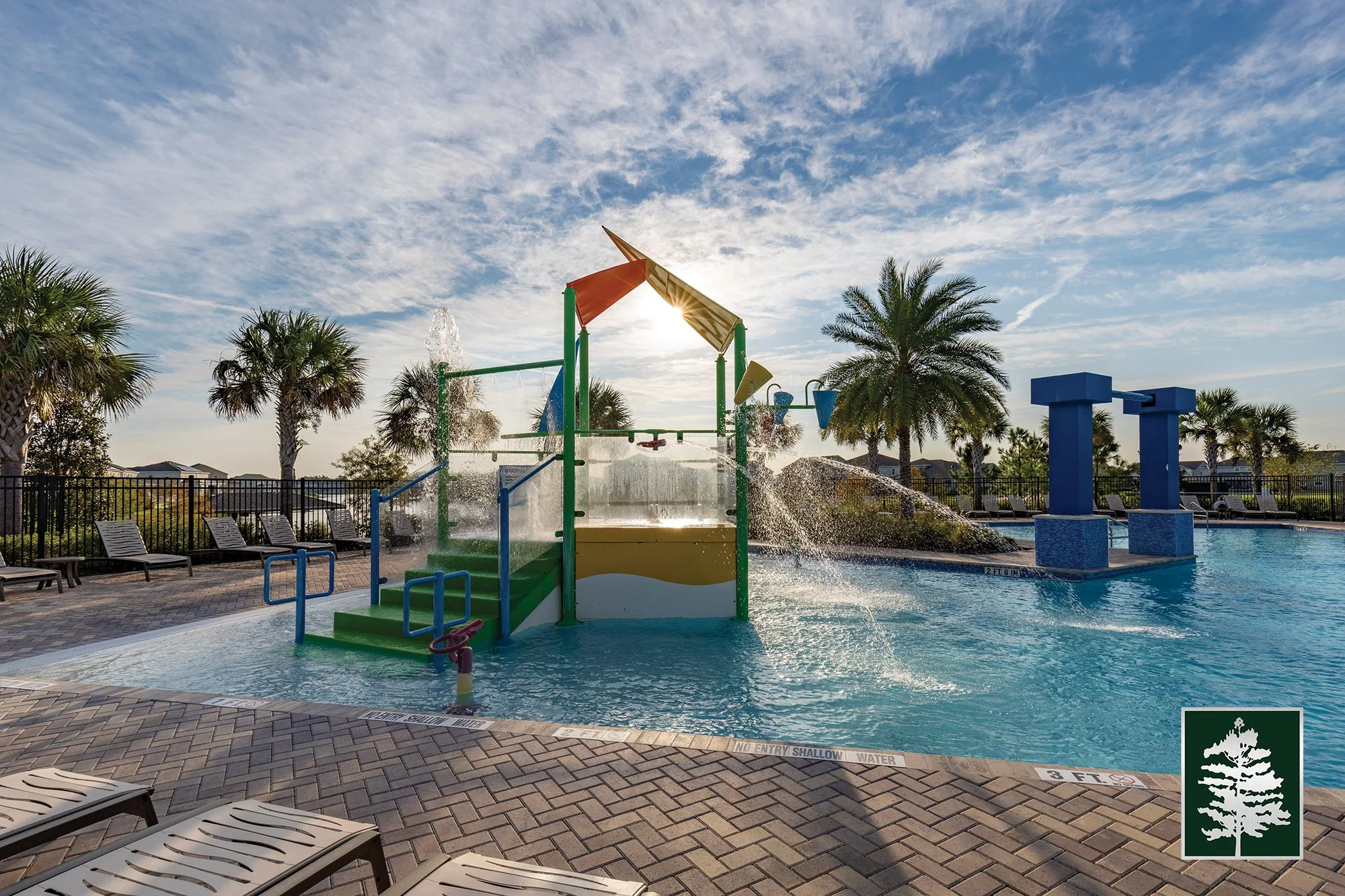 A colorful water play area with a small water fountain or structure in a pool, surrounded by palm trees, lounge chairs, and a safety fence, under a partly cloudy sky.
