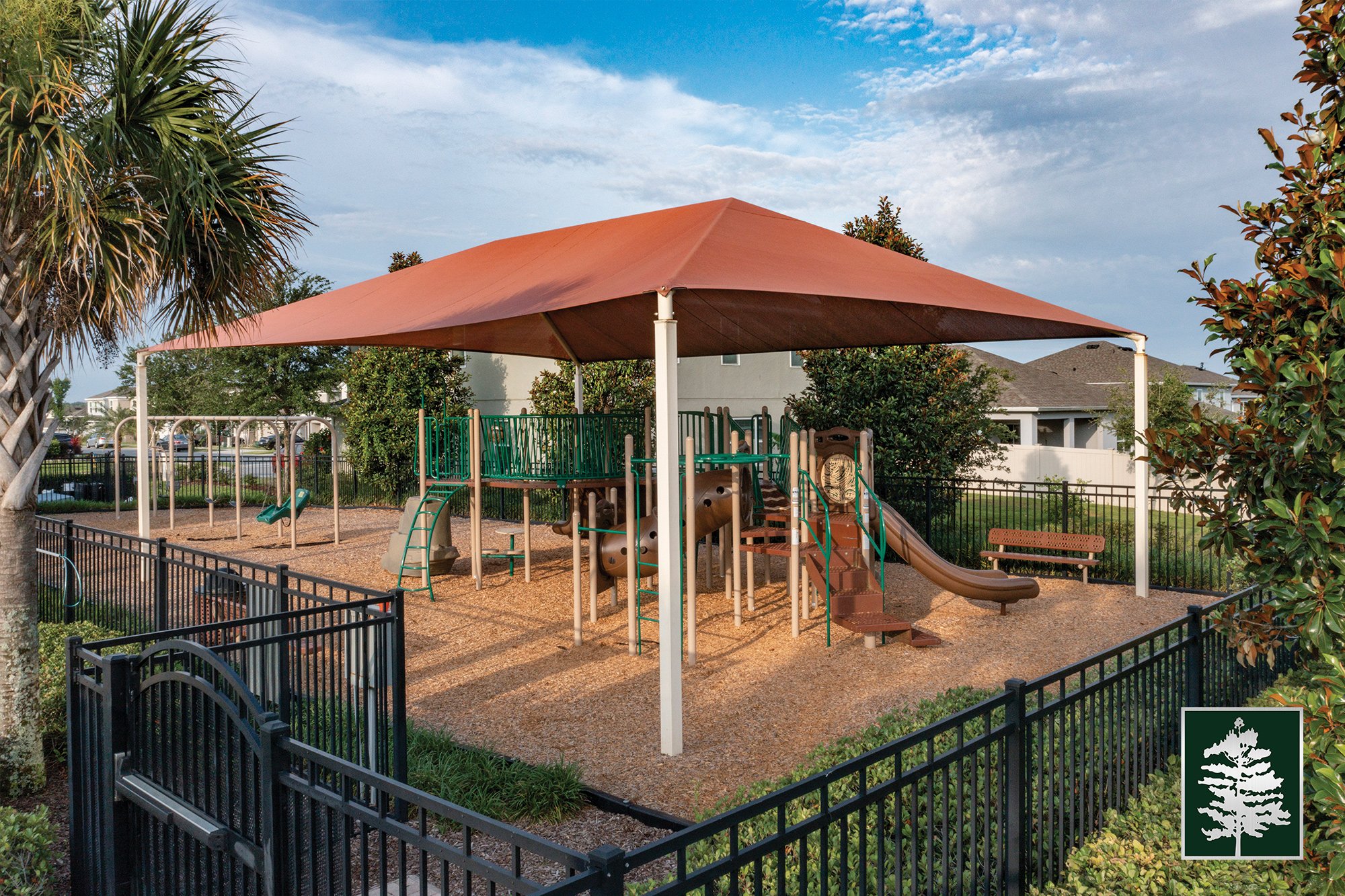 Playground with slides, climbing structures, and swings enclosed by black fence, covered by a large canopy, surrounded by trees and residential buildings in the background.