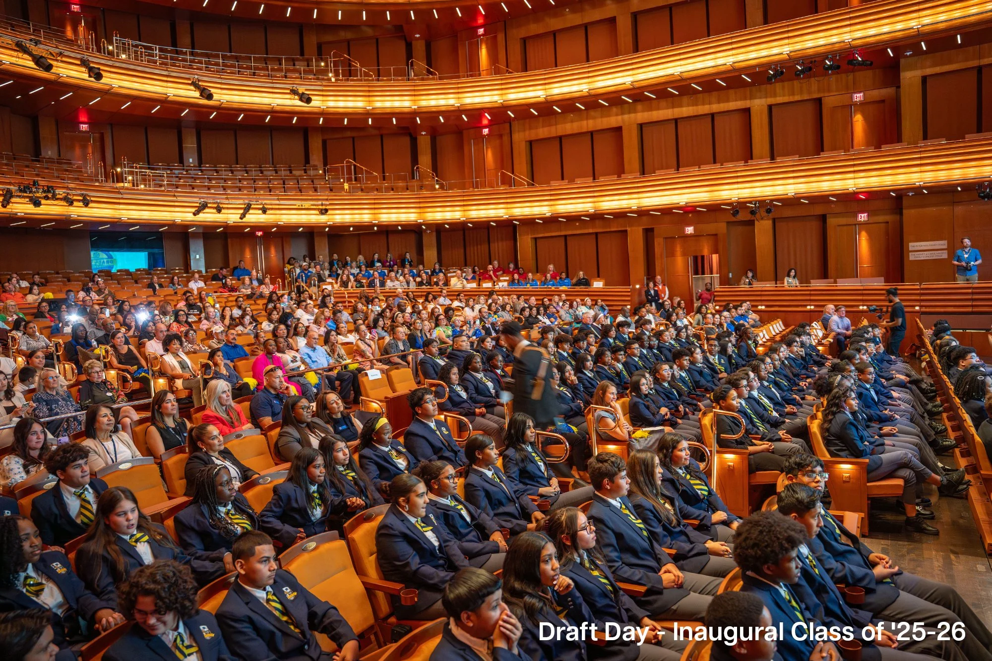 Students in navy blue blazers and striped ties seated in an auditorium for a draft day event of the Inaugural Class of '25-26.