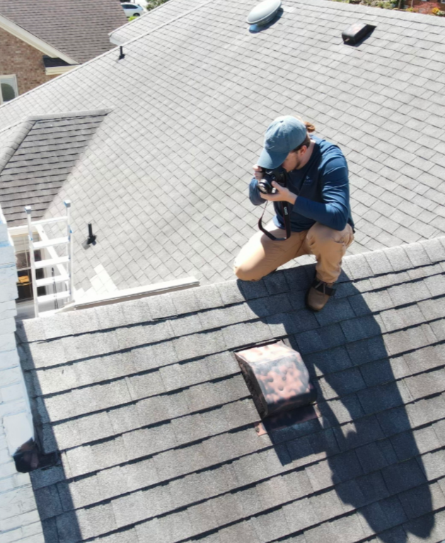 A person kneeling on a house roof, taking a photo with a camera. The roof has gray shingles, and the person is wearing a blue hoodie, beige pants, and a blue cap.
