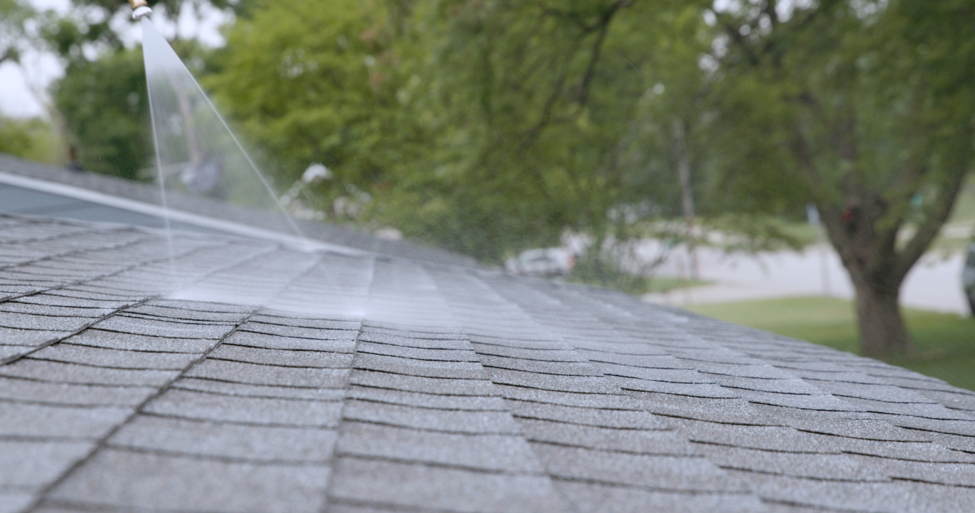 Close-up of a house roof with water spraying from a nozzle, with trees and a street in the background.