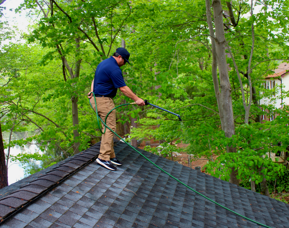 A man wearing a navy shirt, tan pants, and a baseball cap is on a sloped roof, using a long pole with a scrub brush attached to clean the shingles, surrounded by green trees.