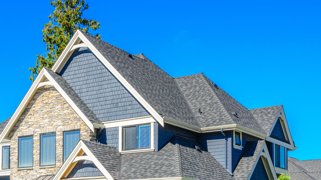 Close-up of a large house's roof with a mix of gray shingles and stone siding, under a blue sky with a pine tree in the background.
