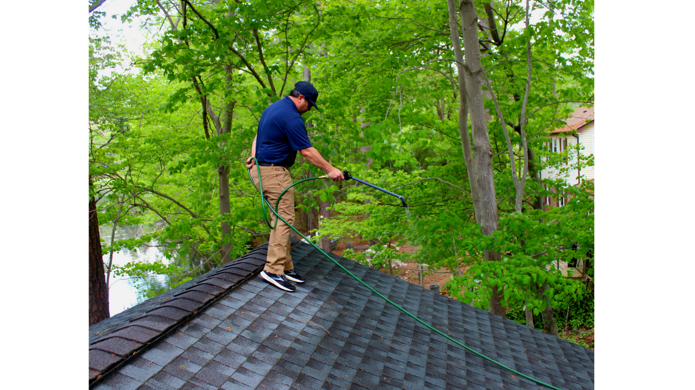 A man wearing a blue shirt, khaki pants, and a blue cap is standing on a sloped roof and using a pressure washer to clean it. He is surrounded by green trees.