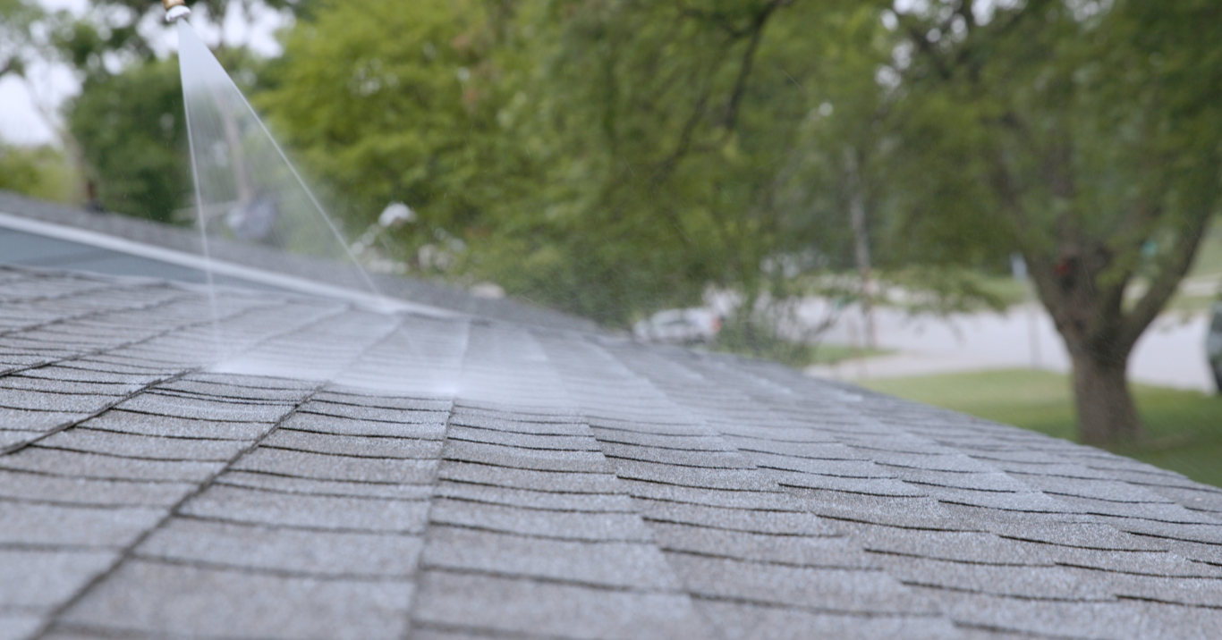 A house roof with an installed sprinkling system spraying water onto the shingles, surrounded by trees and a residential neighborhood.
