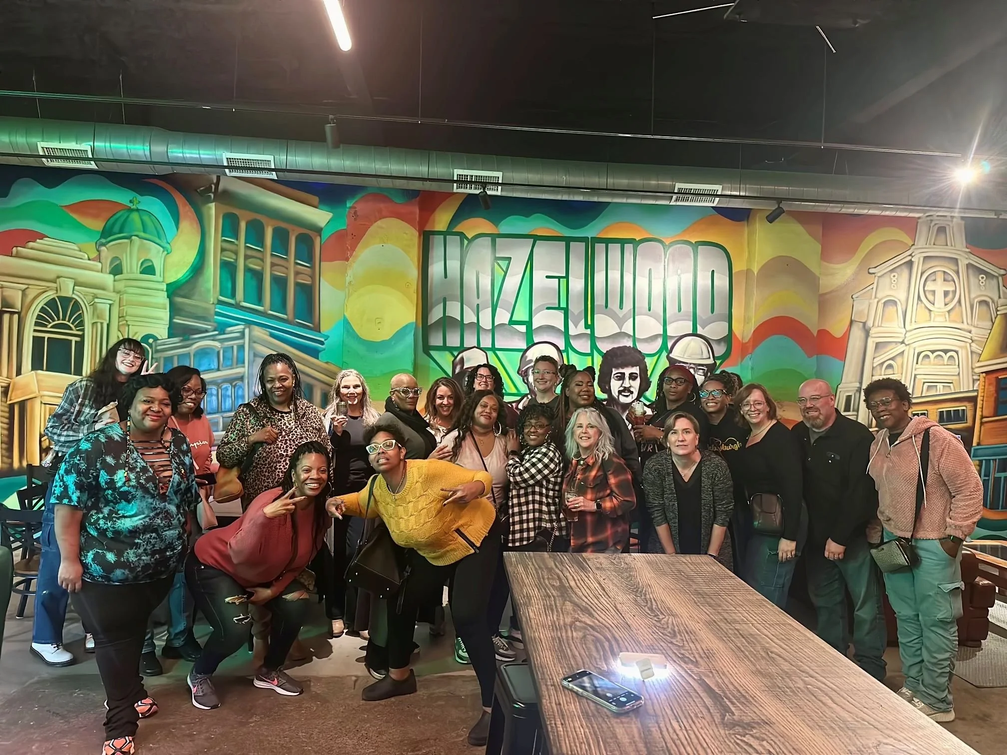 Group of diverse people posing for a photo inside the Hazel Grove Brewing taproom with the colorful mural art of Hazelwood in the background.