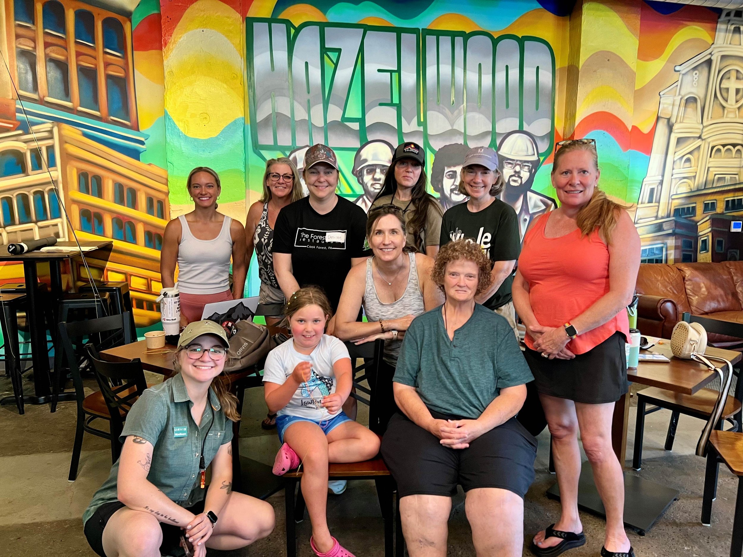 A group of eleven women and two children posing inside the Hazel Grove Brewing taproom with a vibrant mural featuring abstract buildings and portraits of steel workers in background.