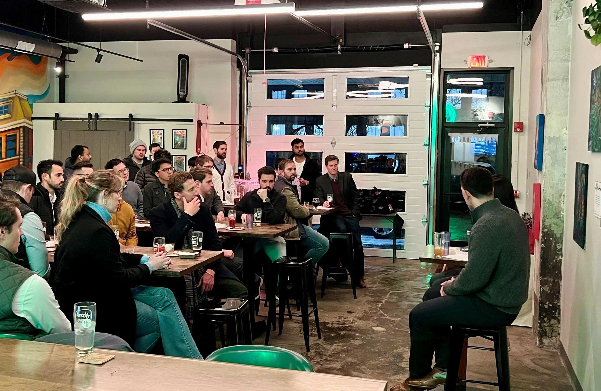 A group of people attending a presentation in the Hazel Grove Brewing taproom.  The audience is seated on chairs and standing, listening attentively while holding glasses of craft beer.